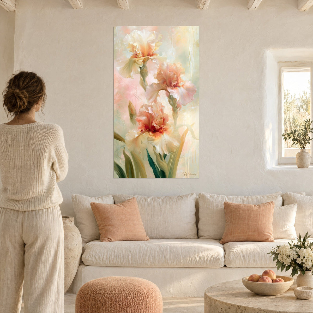 Quadro con tre iris avorio e pesca dai petali ondulati, sfondo rosa cipria, donna di spalle, sopra un divano in un soggiorno luminoso