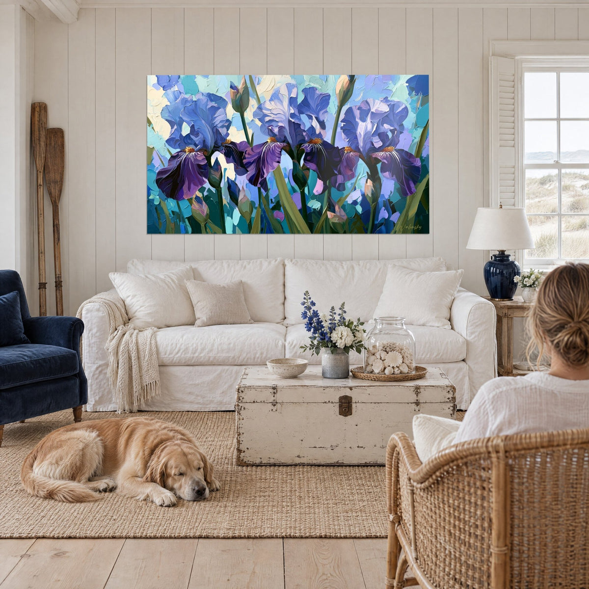 Quadro con tre iris viola e lavanda in pittura densa, foglie verde oliva, sopra un divano in un soggiorno luminoso
