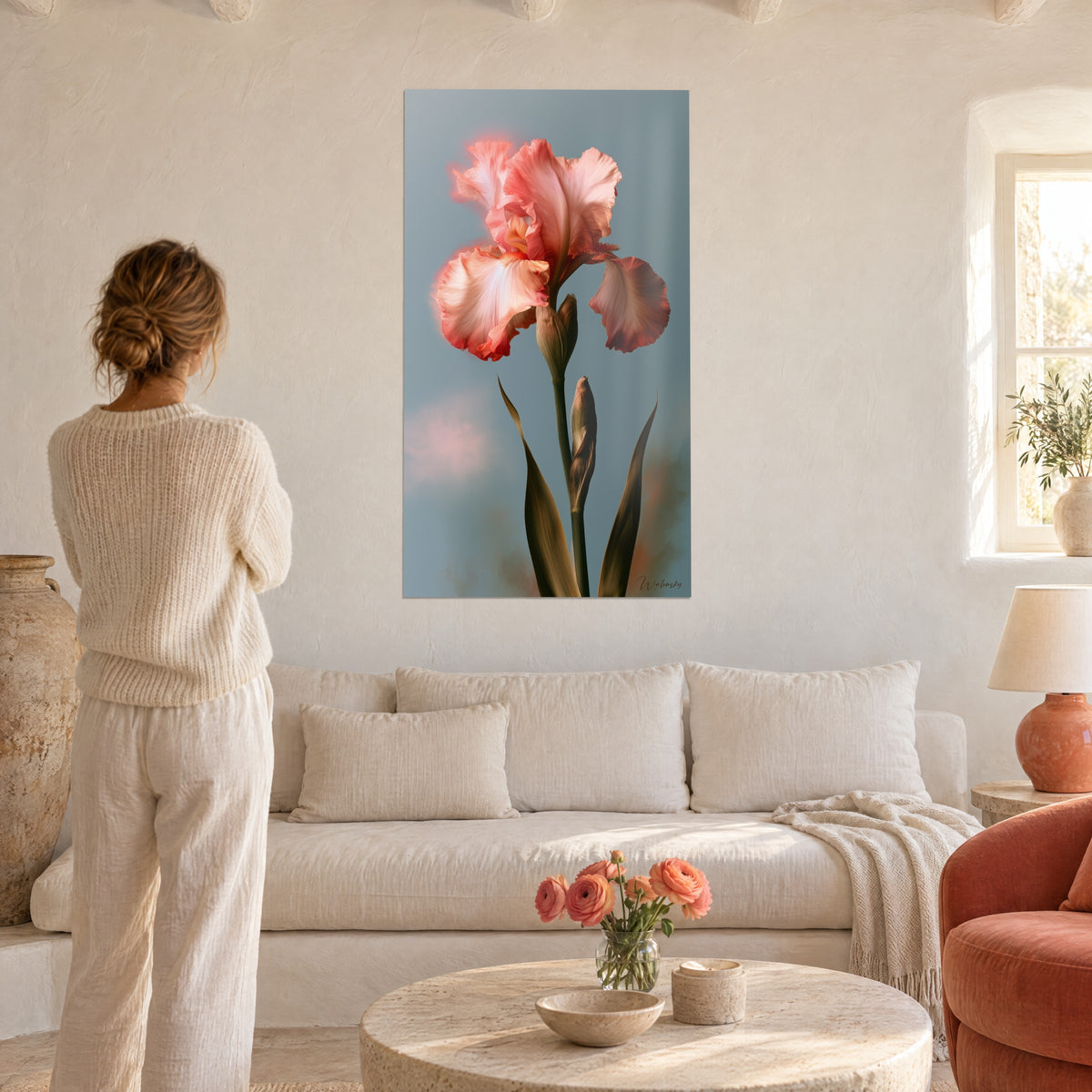 Schilderij iris in koraalroze met golvende bloemblaadjes op grijsblauwe achtergrond, vrouw van achteren in een crèmekleurige trui, boven een bank in een lichte woonkamer