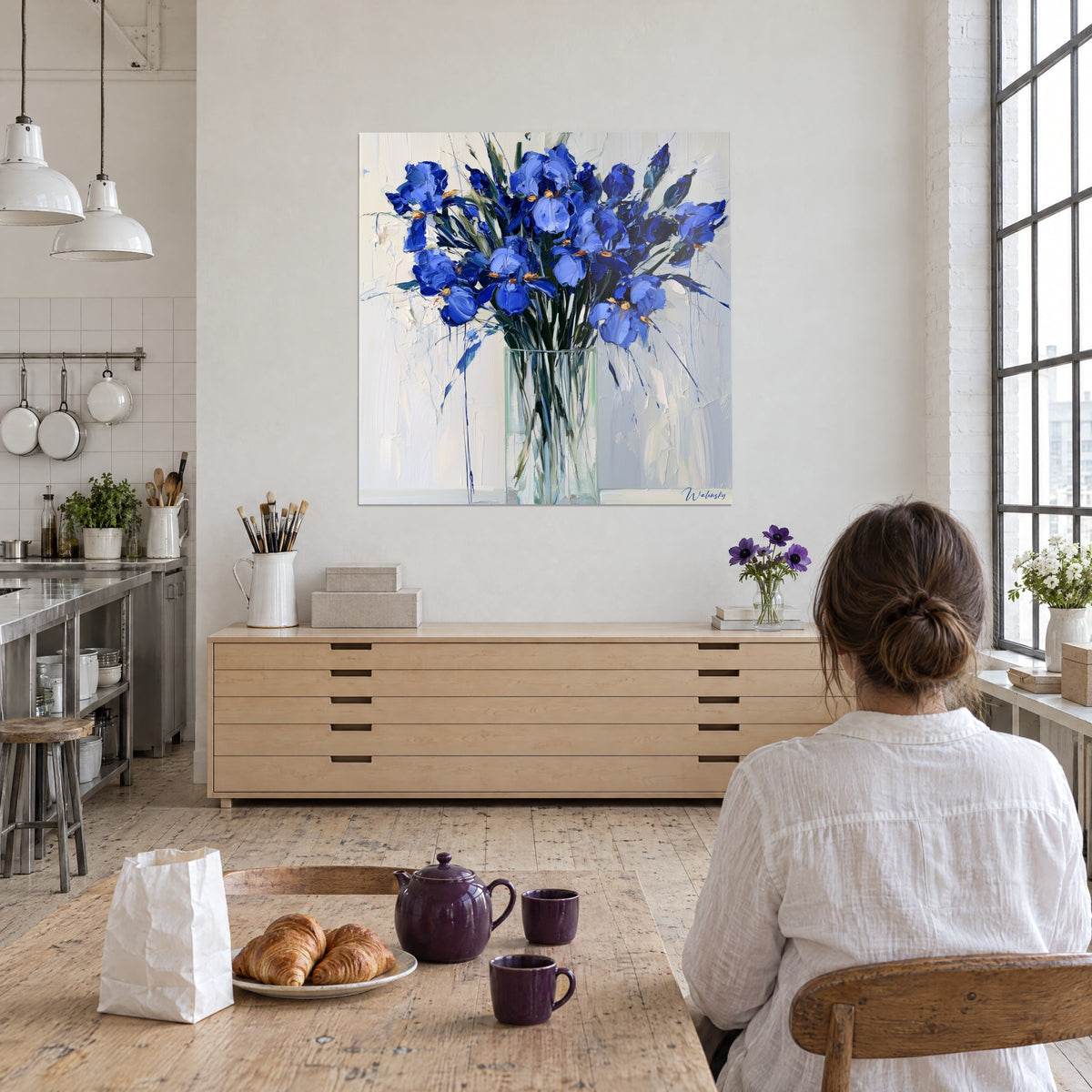 Cuadro iris morados en un jarrón transparente, pinceladas de espátula azules y marfil, mujer de espaldas sentada a una mesa con croissants, loft de artista