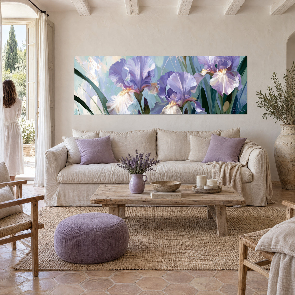 Wall art with purple irises and crumpled petals on a frosted blue background, a woman seen from behind in the foreground, above a sofa in a bright living room
