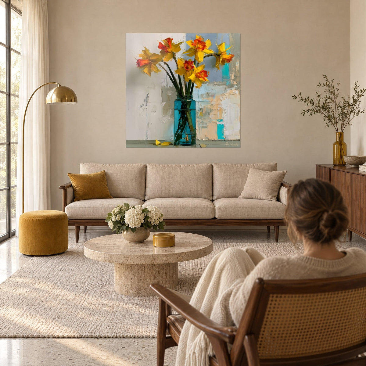 Cuadro de narcisos amarillo sol con corazón anaranjado, jarrón turquesa, fondo gris azulado, mujer de espaldas, encima de un sofá en un salón luminoso