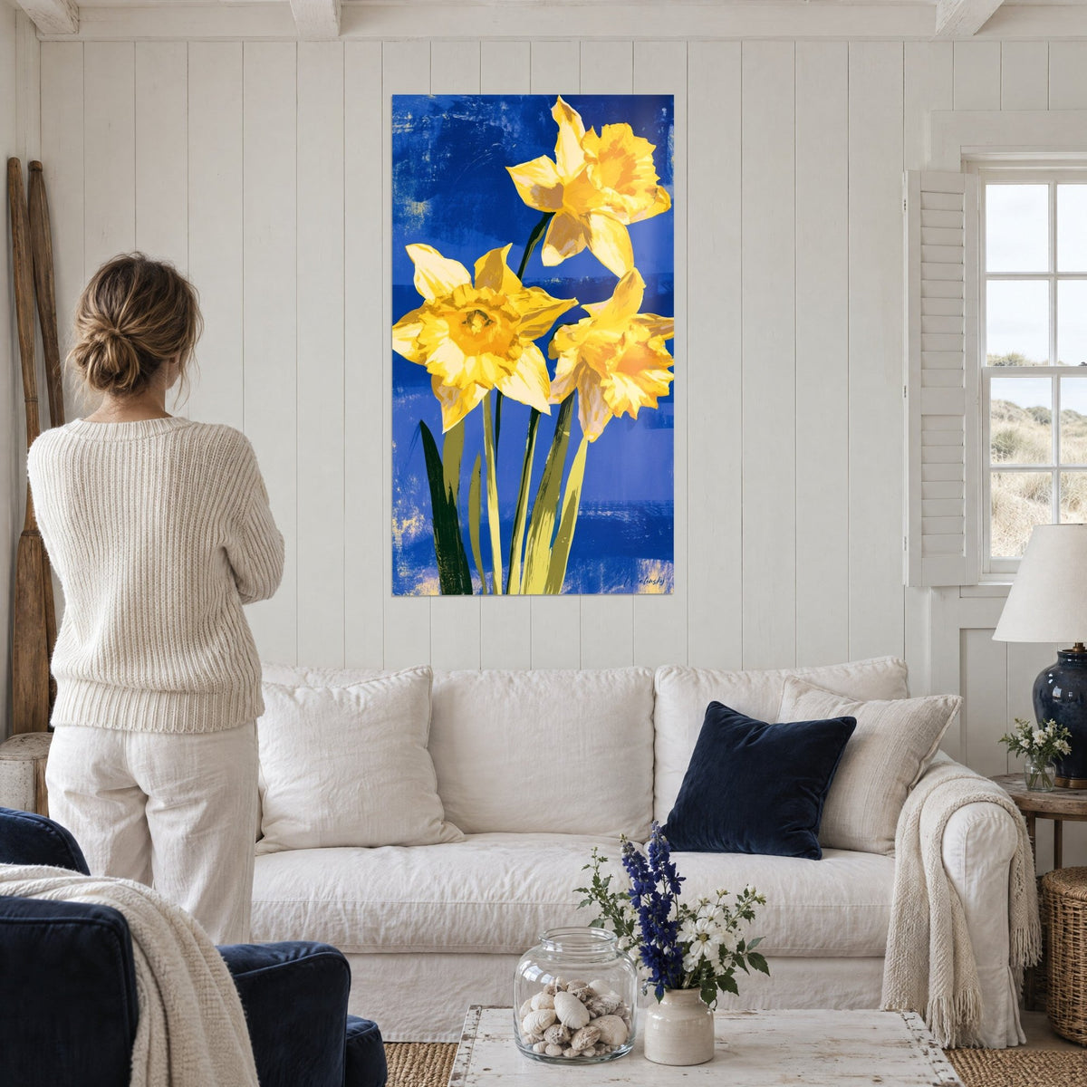 Wall art golden daffodils on royal blue background, a woman seen from behind gazes at it, above a sofa in a bright living room