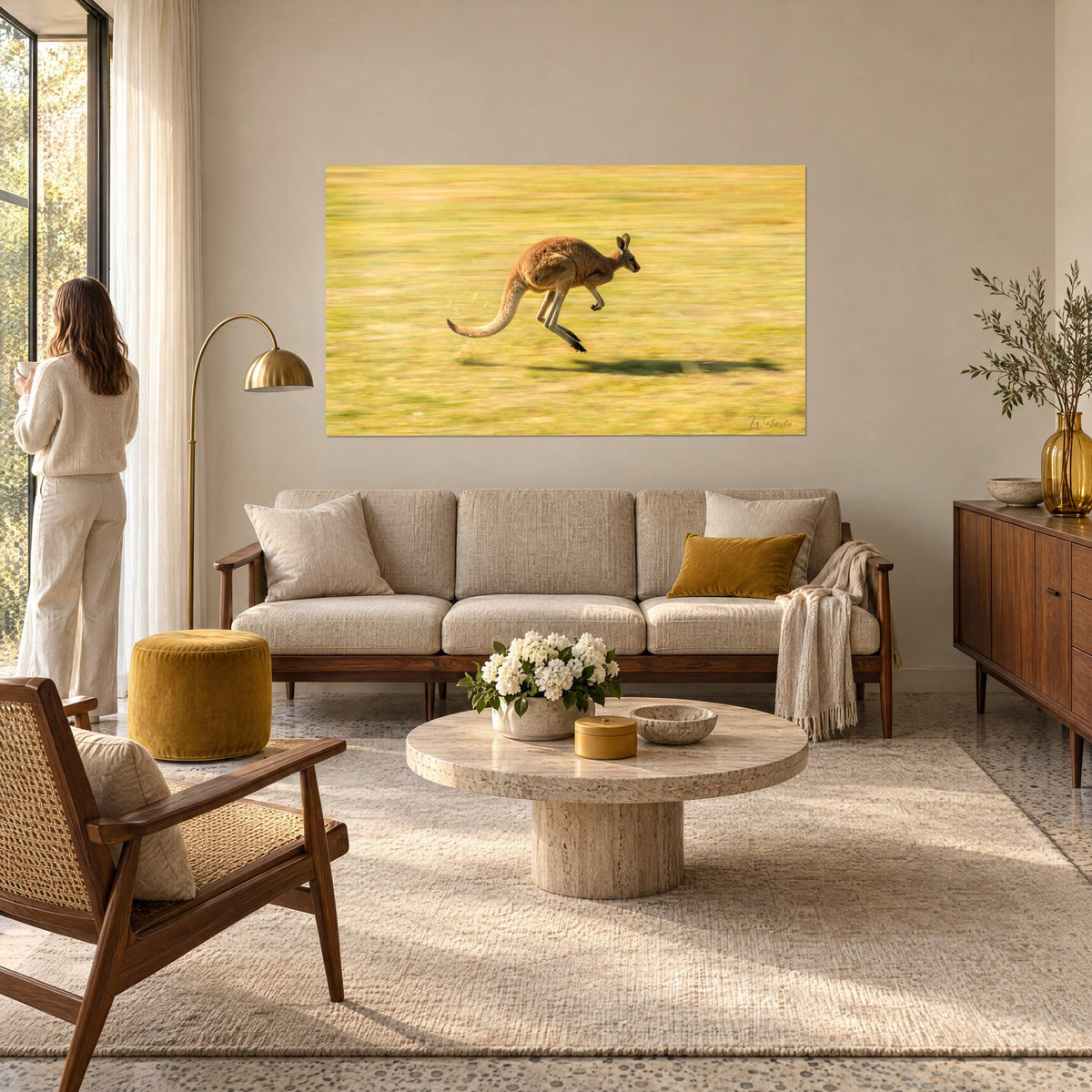Tableau kangourou roux bondissant sur une prairie dorée, ombre allongée au sol, femme de dos, au-dessus d'un canapé dans un salon lumineux