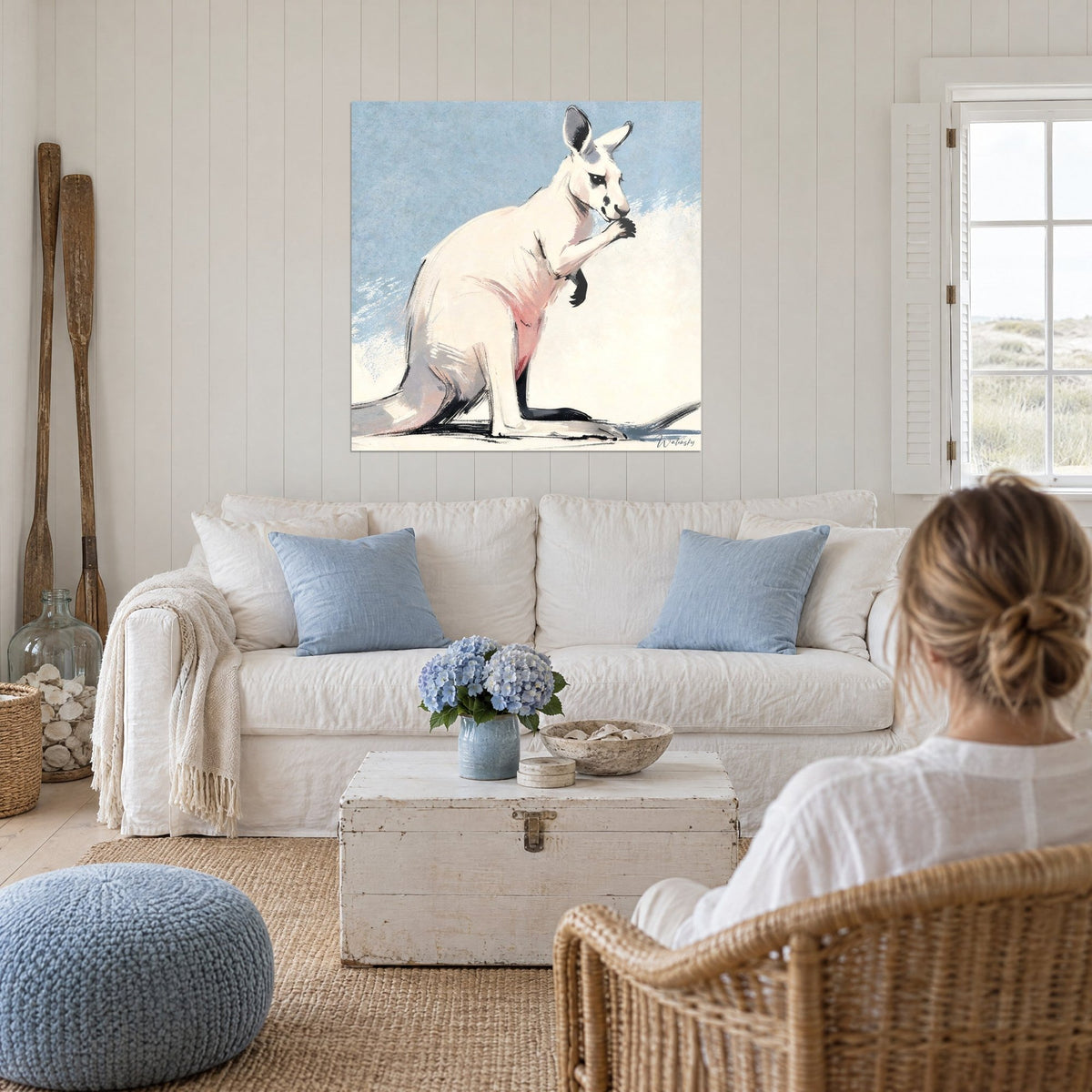 Tableau kangourou blanc aux flancs rosés grignotant sa patte, fond bleu ciel, au-dessus d'un canapé dans un salon lumineux