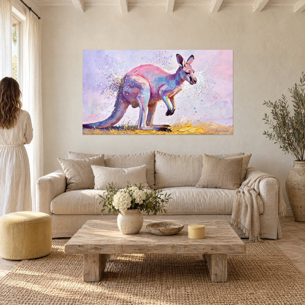 Cuadro marsupial rosa y azul salpicado de gotas de colores, fondo lila, una mujer de espaldas, encima de un sofá en un salón luminoso