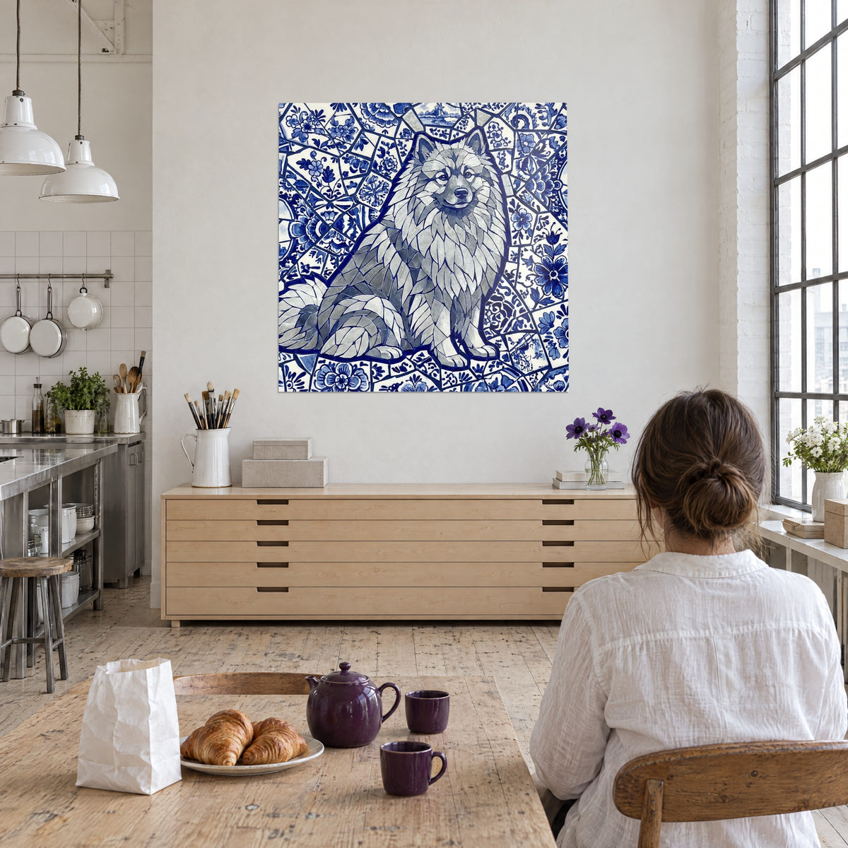 Tableau loulou à poil clair en mosaïque de Delft, bleu marine et blanc, regardé par une femme de dos attablée devant des croissants