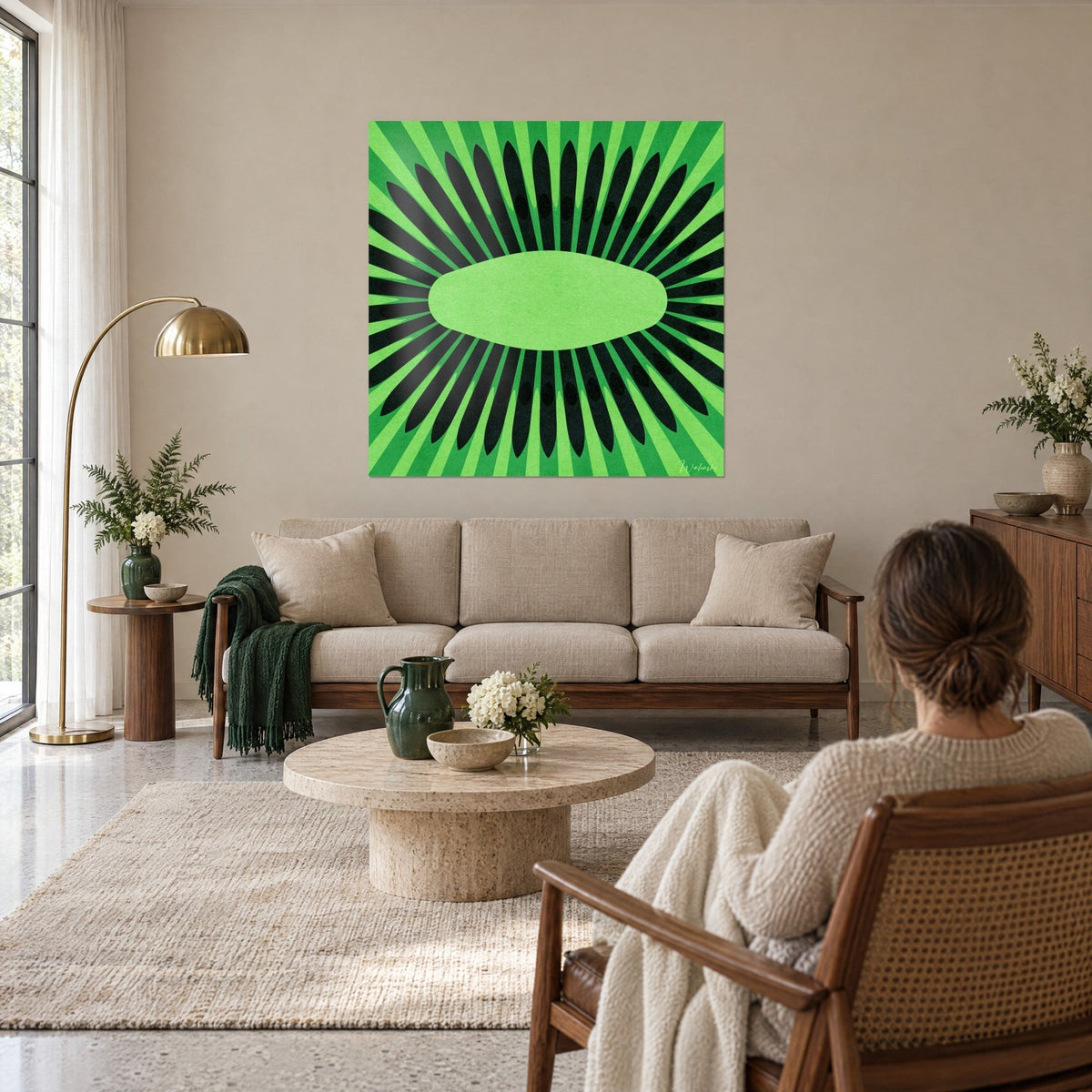 Wall art kiwi cut in half, black seeds radiating around a soft green heart, woman seen from behind, above a sofa in a bright living room