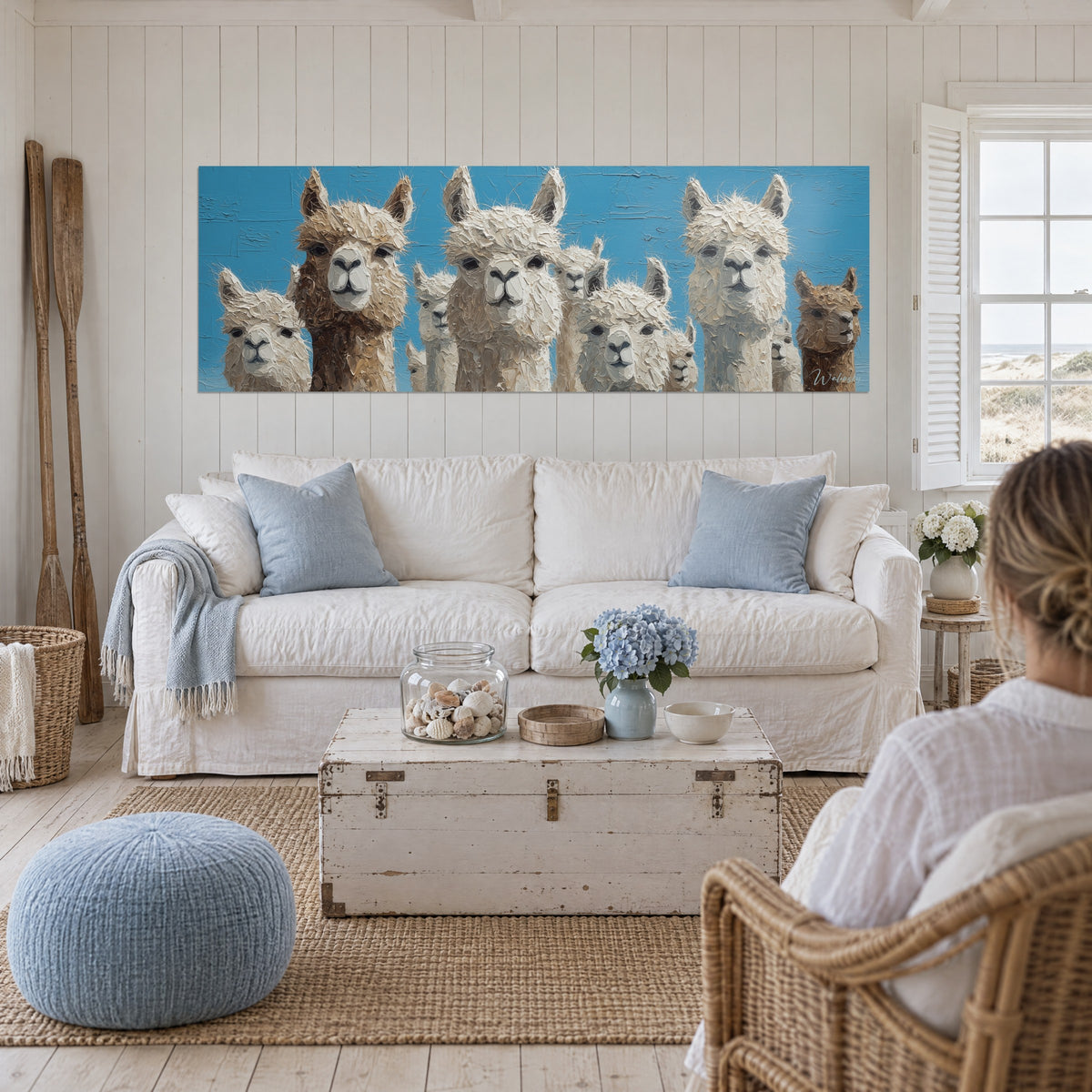 Cuadro rebaño de llamas lanudas, pelaje crema y marrón sobre cielo azul, encima de un sofá en un salón luminoso
