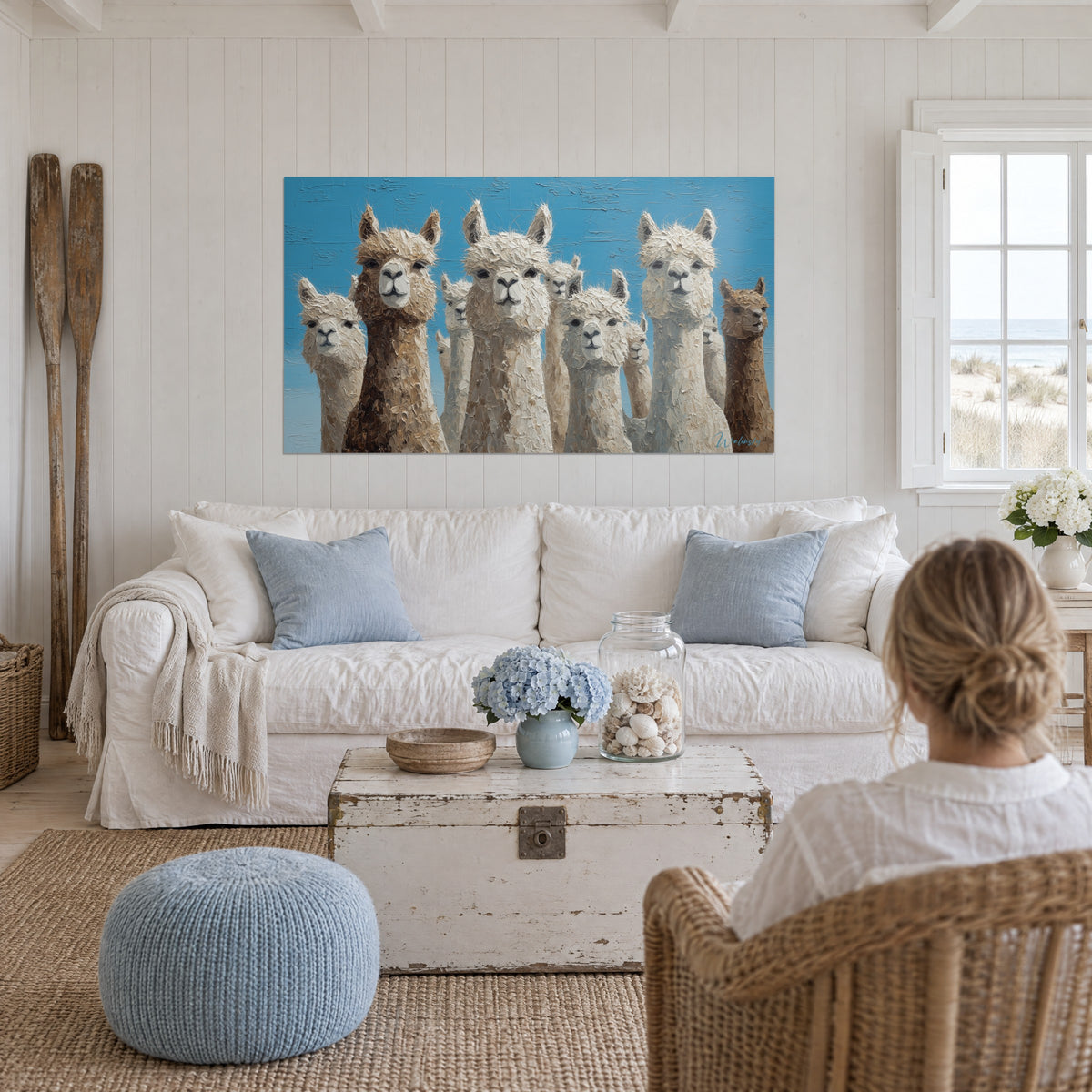 Wall art herd of llamas with cream and caramel wool against a blue sky, woman seen from behind, above a sofa in a bright living room