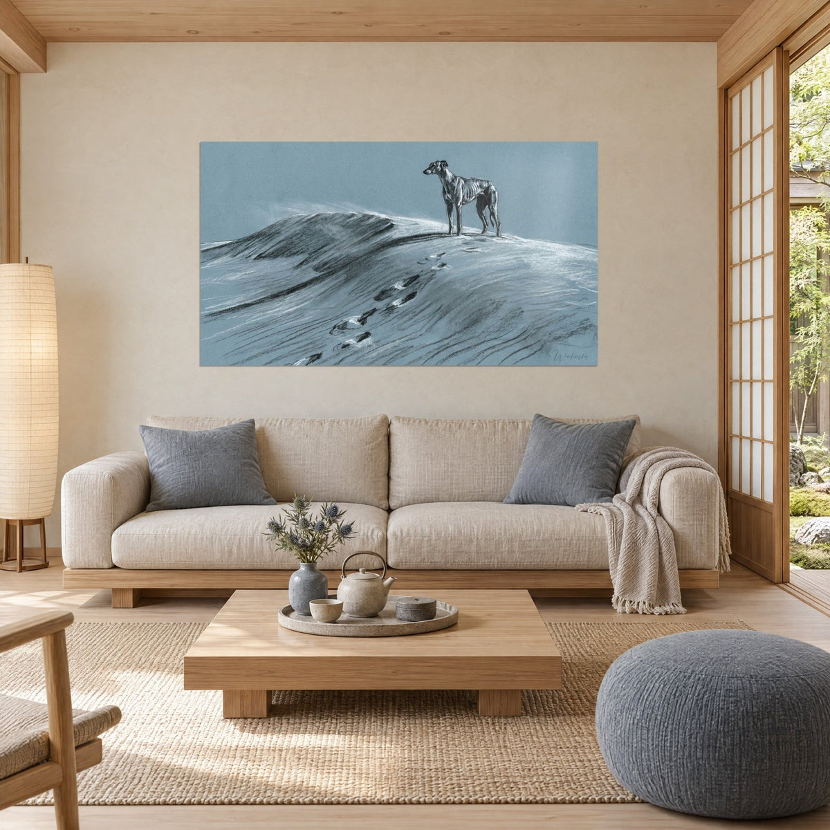 Wall art of a slender greyhound standing on a snowy dune, black charcoal and white chalk on a blue-grey background, above a sofa in a bright living room