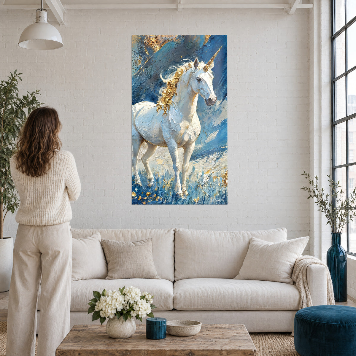 Tableau licorne blanche à corne dorée, crinière en feuilles d'or sur fond bleu, femme de dos au-dessus d'un canapé dans un salon lumineux