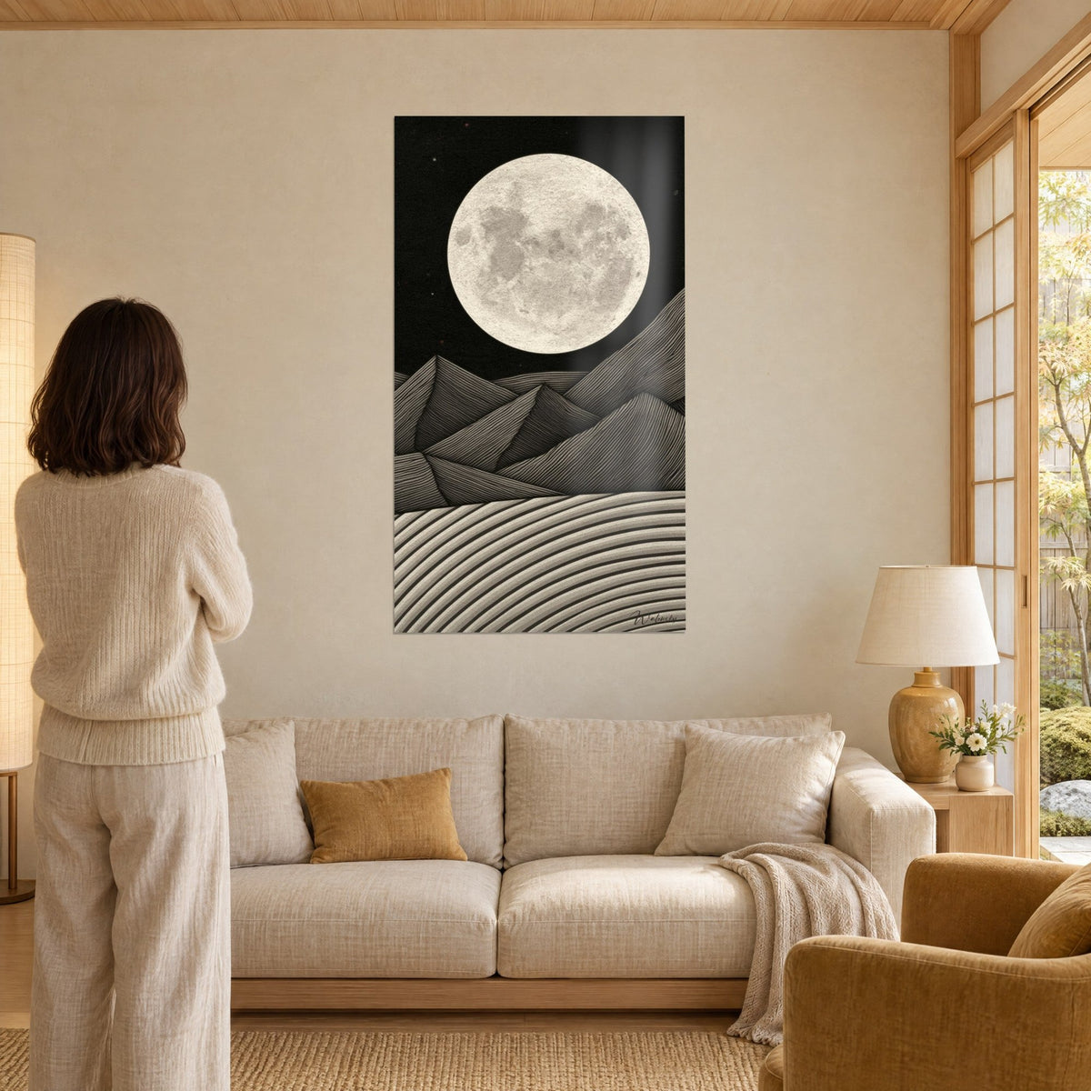 Wall art with a textured full moon on a black sky, grey and ivory striped mountains, woman seen from behind, above a sofa in a bright living room