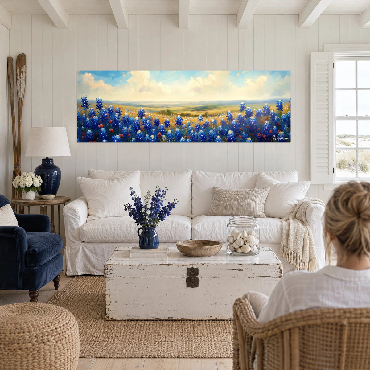 Schilderij veld met blauwe lupinen onder een crème en azuurblauwe lucht, gouden vallei in de verte, twee personen van achteren boven een bank in een lichte woonkamer