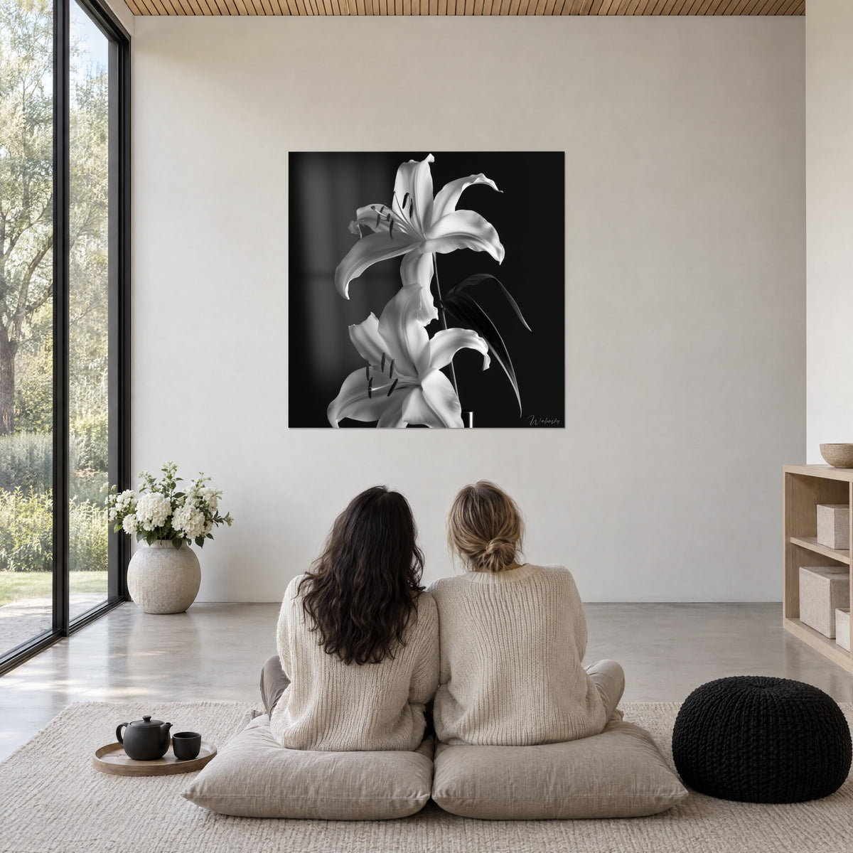 Cuadro de un ramo de dos lirios blancos con estambres negros sobre fondo oscuro, dos mujeres sentadas de espaldas en cojines grises junto a un ventanal