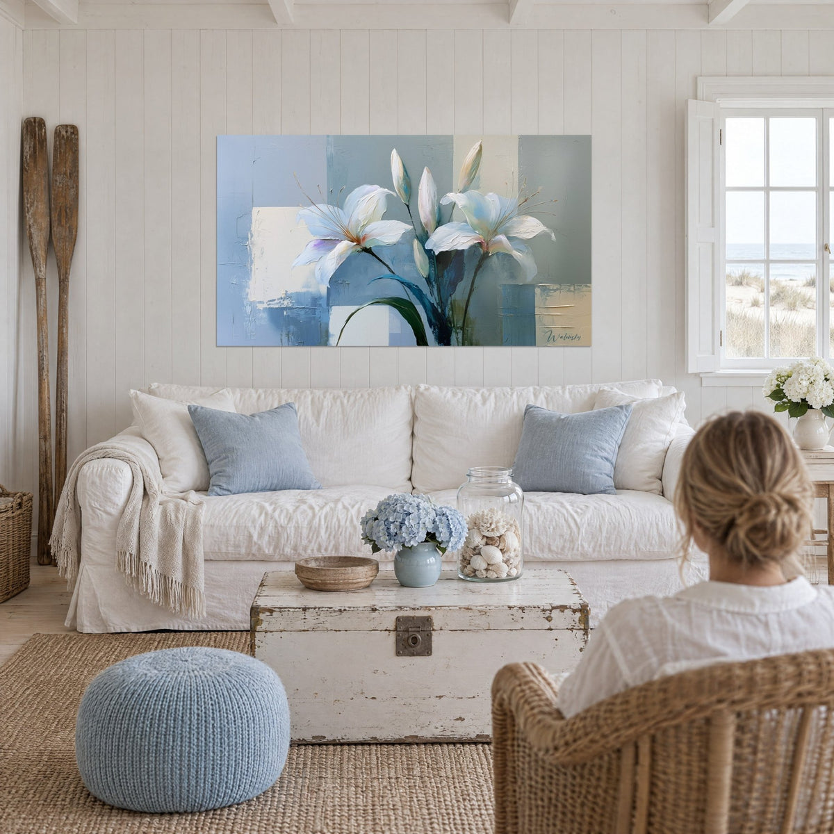 Tableau lys blanc aux pétales épais, tiges effilées ocre et fond bleu-gris, au-dessus d'un canapé dans un salon lumineux