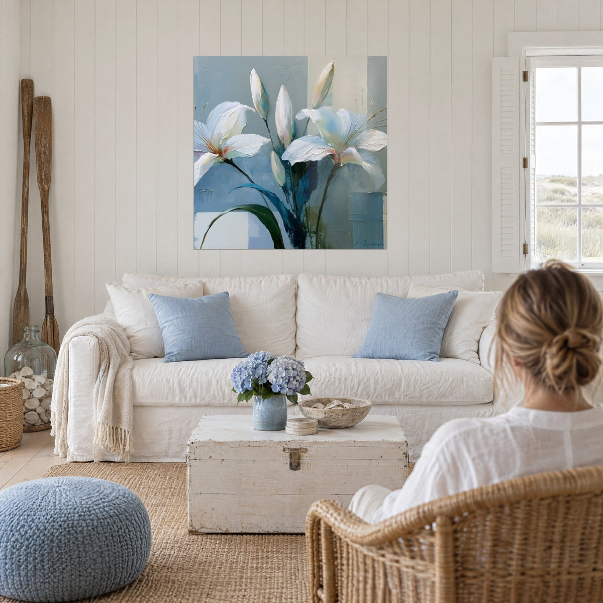 Quadro giglio bianco dai petali madreperlacei e fogliame blu notte, donna di spalle, sopra un divano in un soggiorno luminoso