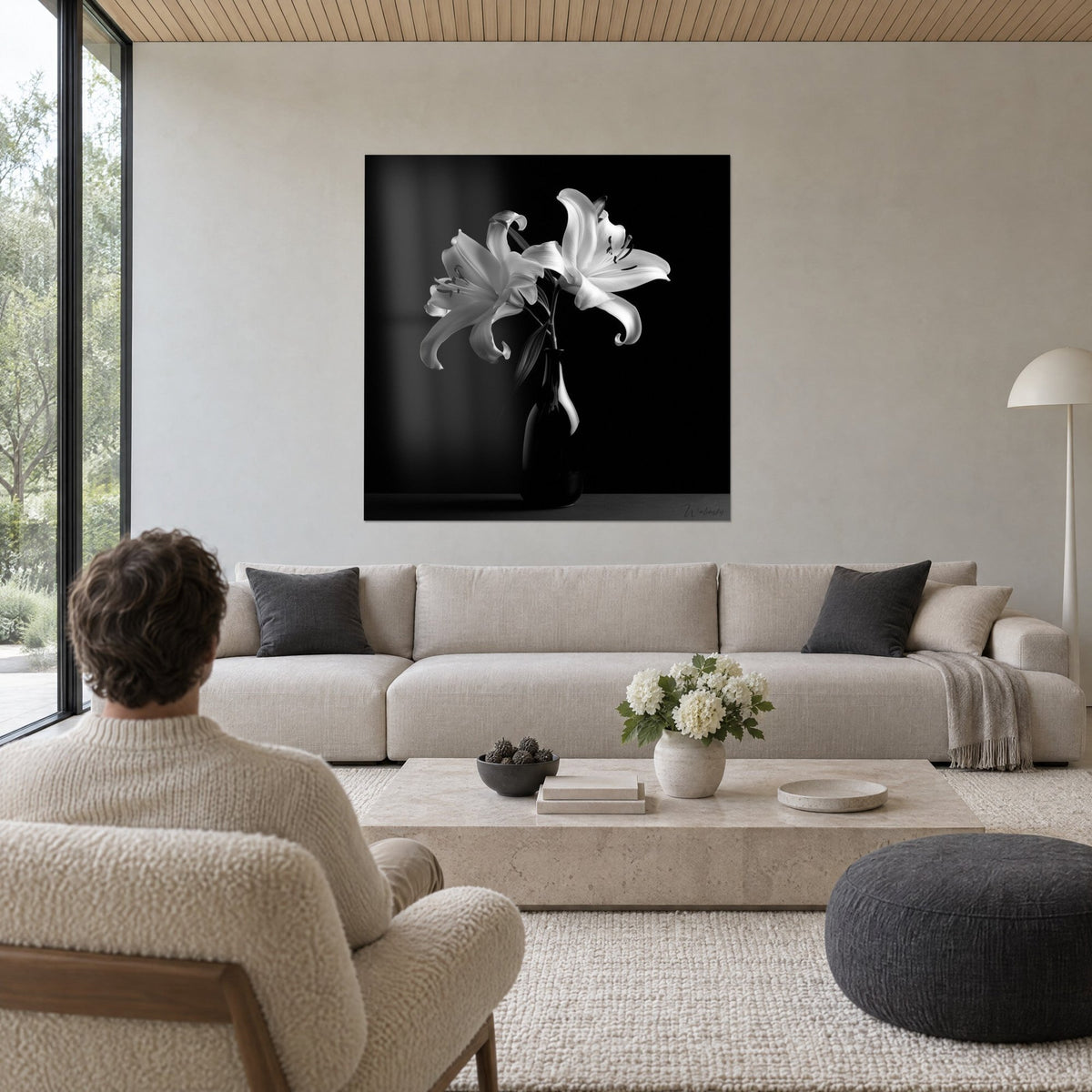 Tableau lys blanc aux pétales recourbés sur fond noir, vase sombre, homme de dos, au-dessus d'un canapé dans un salon lumineux