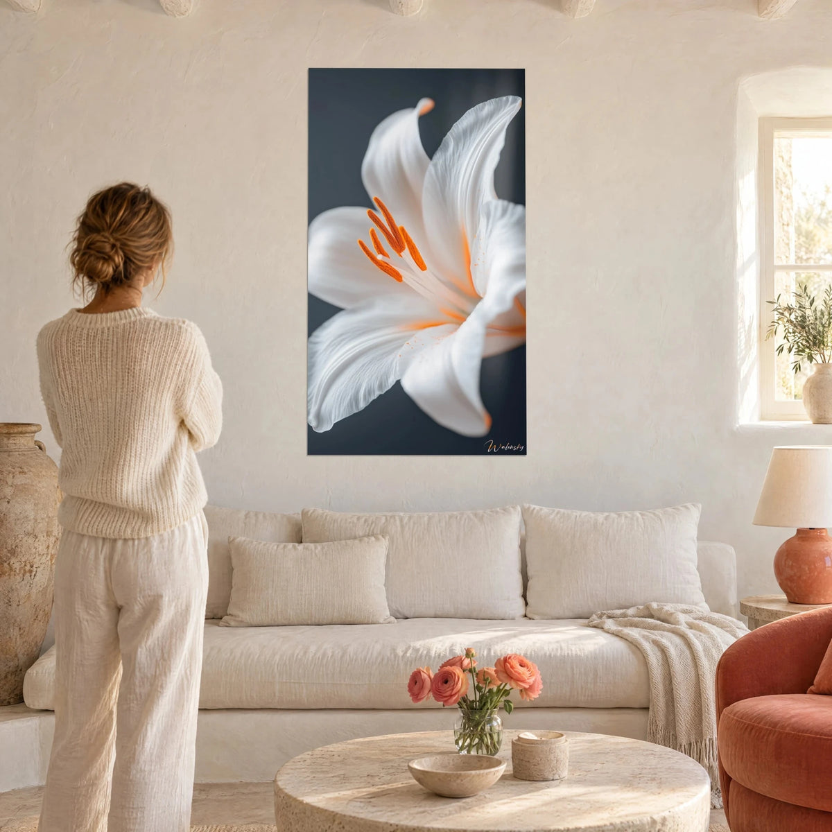 Tableau lys blanc aux étamines orange, fond anthracite, regardé par une femme en pull crème au-dessus d'un canapé dans un salon lumineux