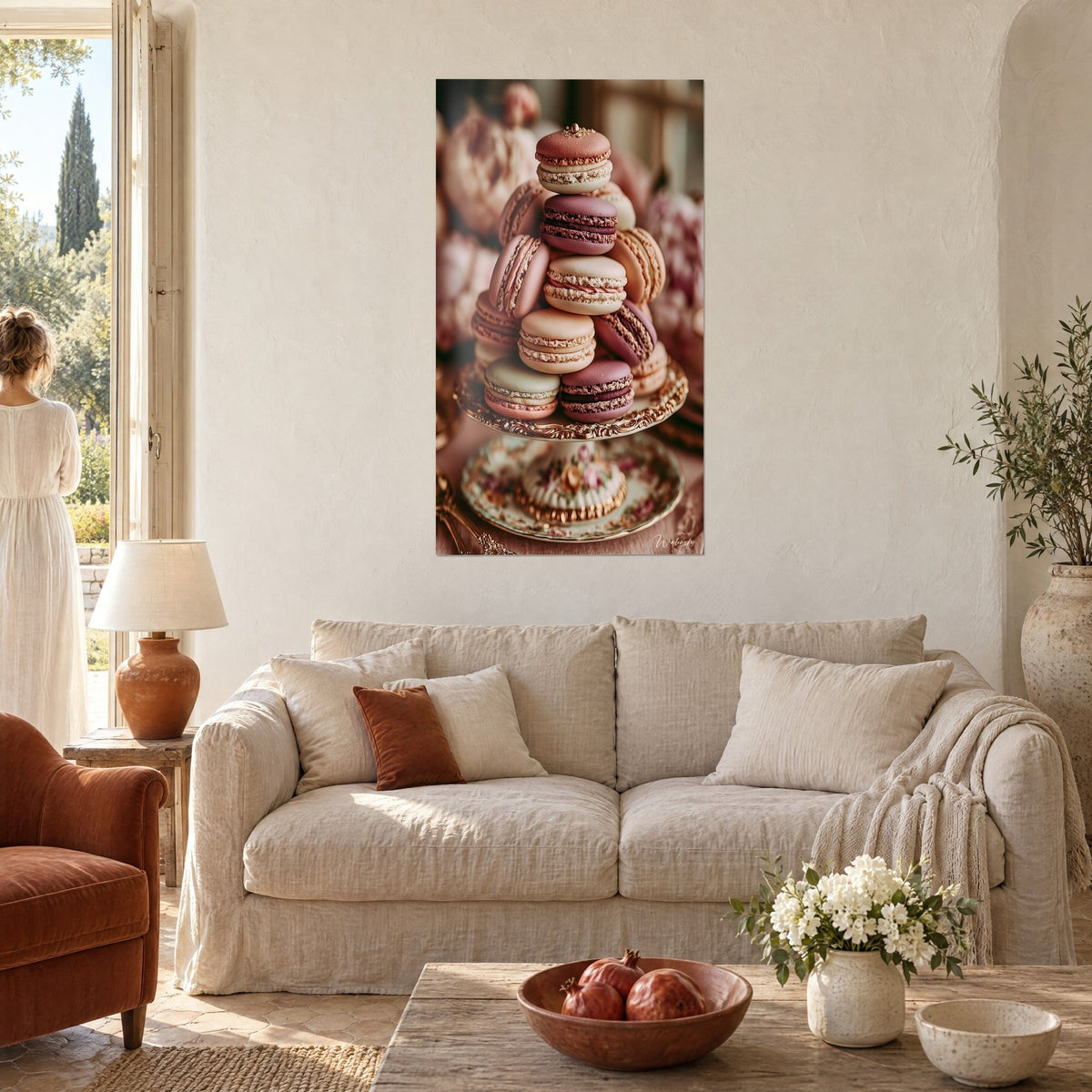 Tableau pyramide de macarons rose poudré, prune et ivoire sur un support doré, personne de dos, au-dessus d'un canapé dans un salon lumineux