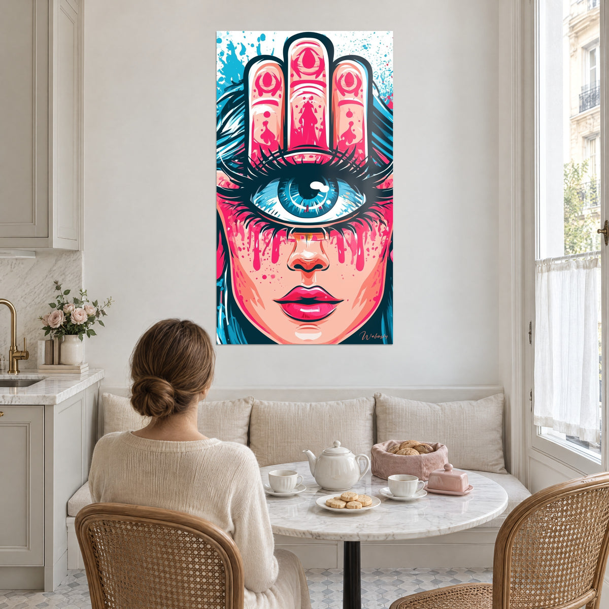 Wall art protective eye in cobalt blue beneath three decorated pink fingers, pink paint tears, facing a woman seated at a table in a bright kitchen