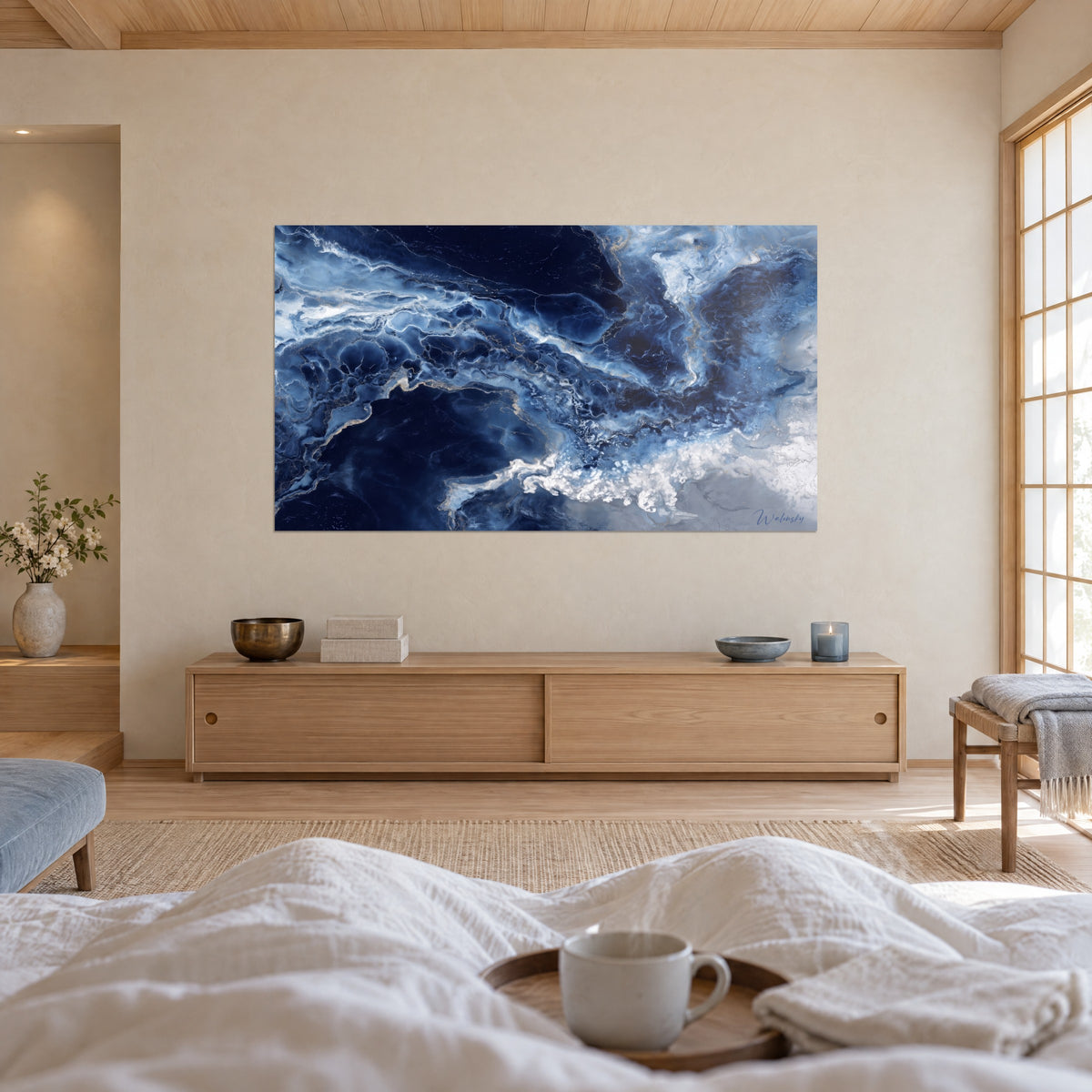 Cuadro formas fluidas con vetas doradas sobre fondo azul marino y blanco roto, persona de espaldas, encima de una cama en un dormitorio claro