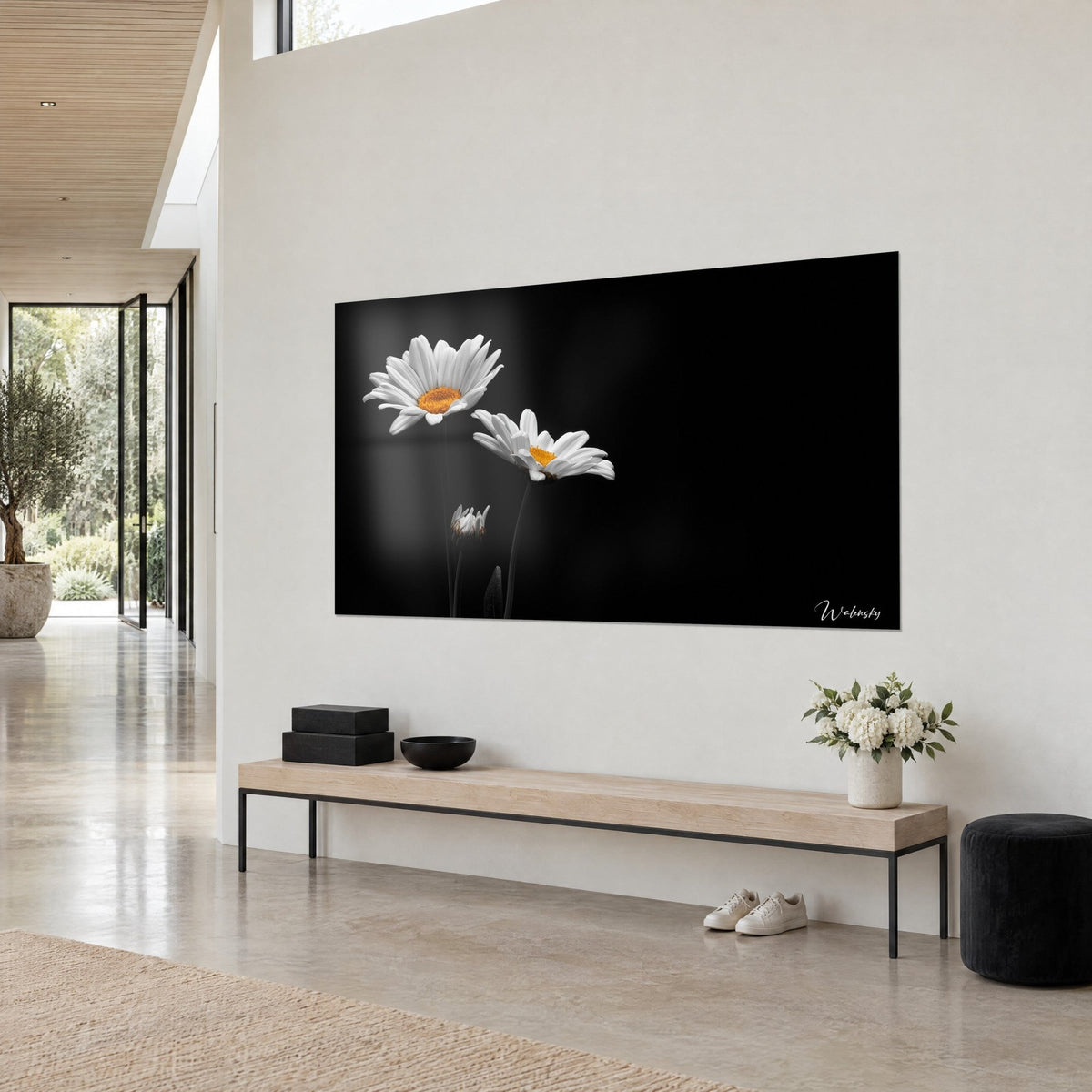 Quadro con fiori di camomilla immacolati e un bocciolo chiuso su fondo antracite, appeso a una parete beige sopra una panca in legno chiaro