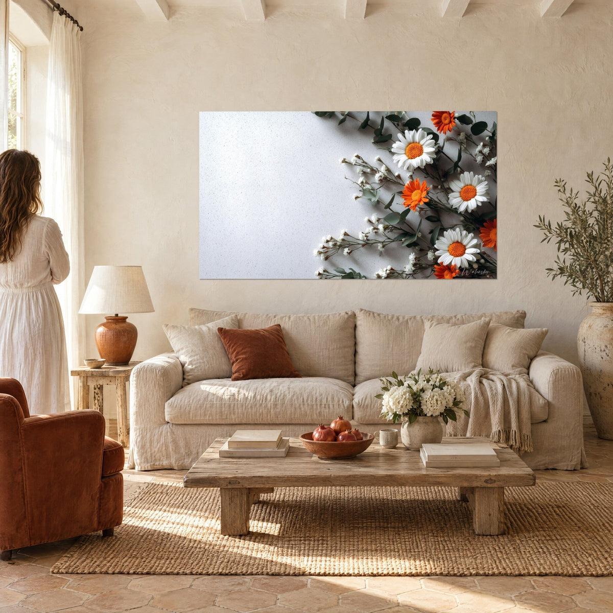 Wall art white daisies with orange centres, grey-green eucalyptus, speckled white background, woman seen from behind above a sofa in a bright living room