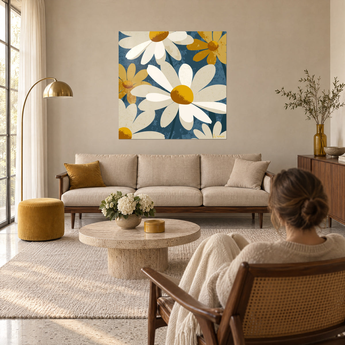 Wall art with ivory daisies and ochre centers on slate blue background, woman seen from behind, above a sofa in a bright living room
