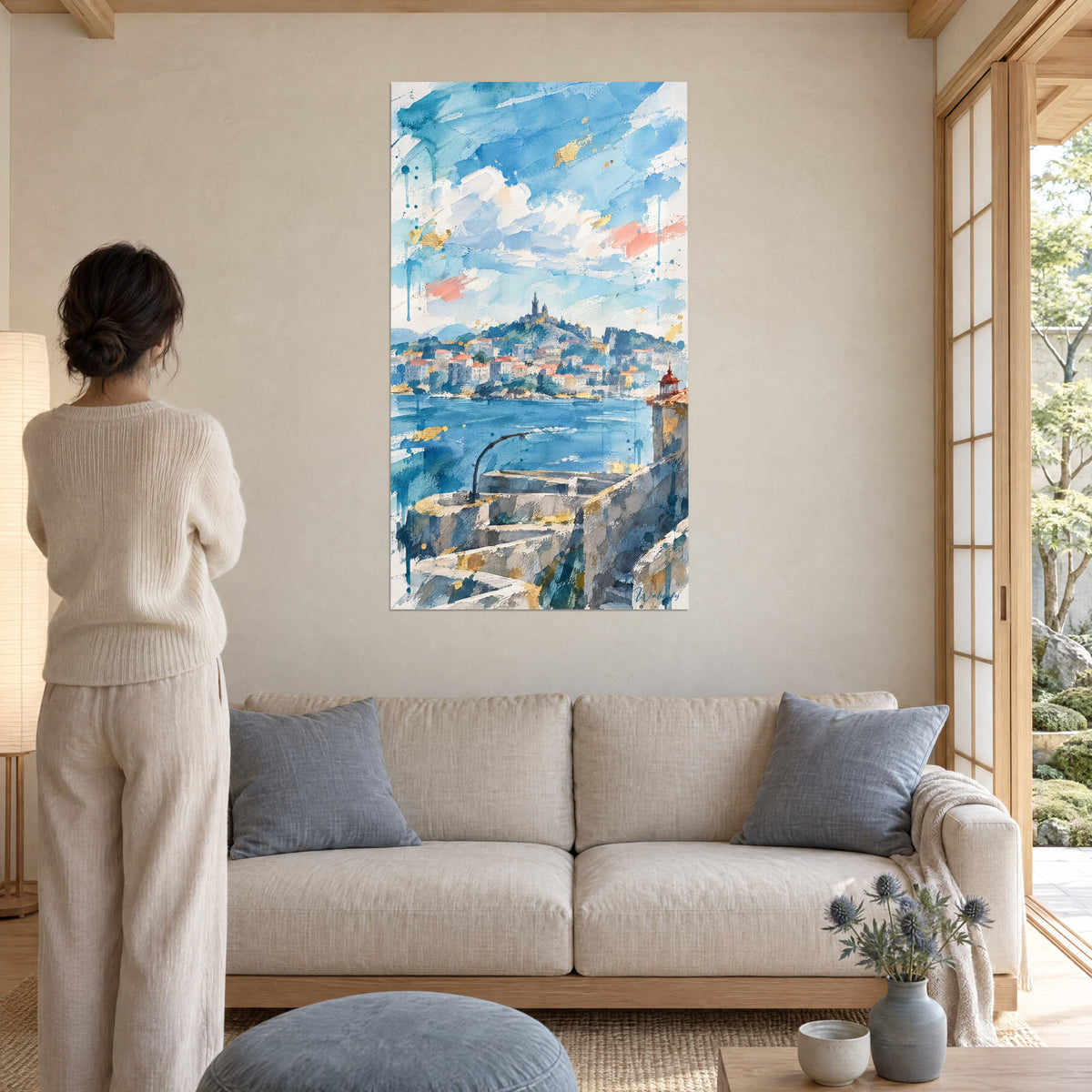 Schilderij basiliek hoog op haar heuvel, hemelsblauwe lucht met koraalkleurige accenten, vrouw van achteren boven een bank in een lichte woonkamer
