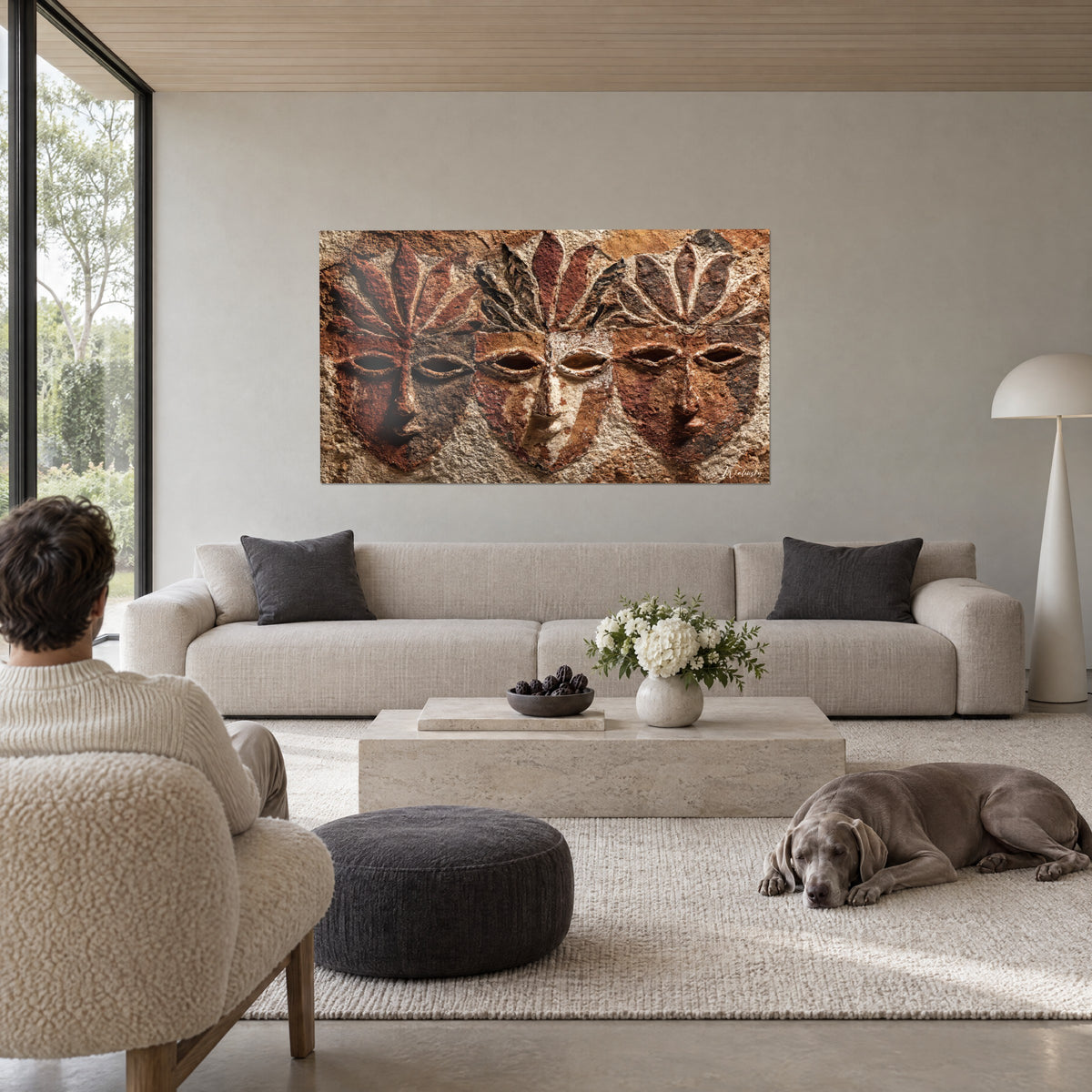 Cuadro tres máscaras esculpidas en relieve, arcilla óxido y blanco crema, coronadas de pétalos, sobre un sofá en un salón luminoso