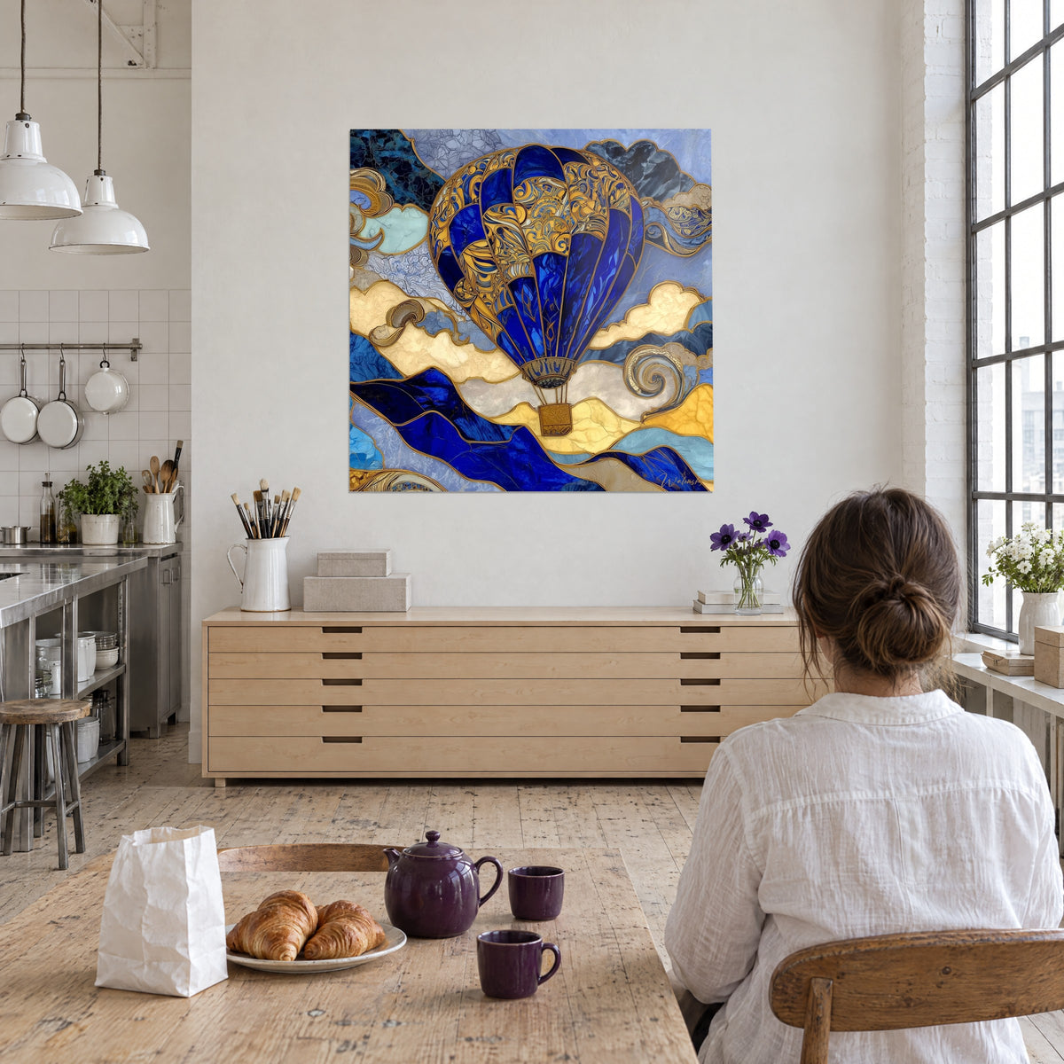 Cuadro globo aerostático en mosaico de vidriera azul real y oro, espirales doradas, frente a una mujer sentada de espaldas a la mesa en un loft