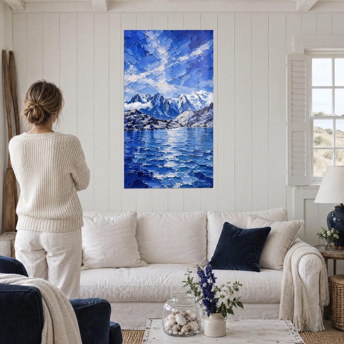 Schilderij van een massief van rotstoppen onder een blauwe en witte hemel, getextureerd meer, boven een bank in een lichte woonkamer
