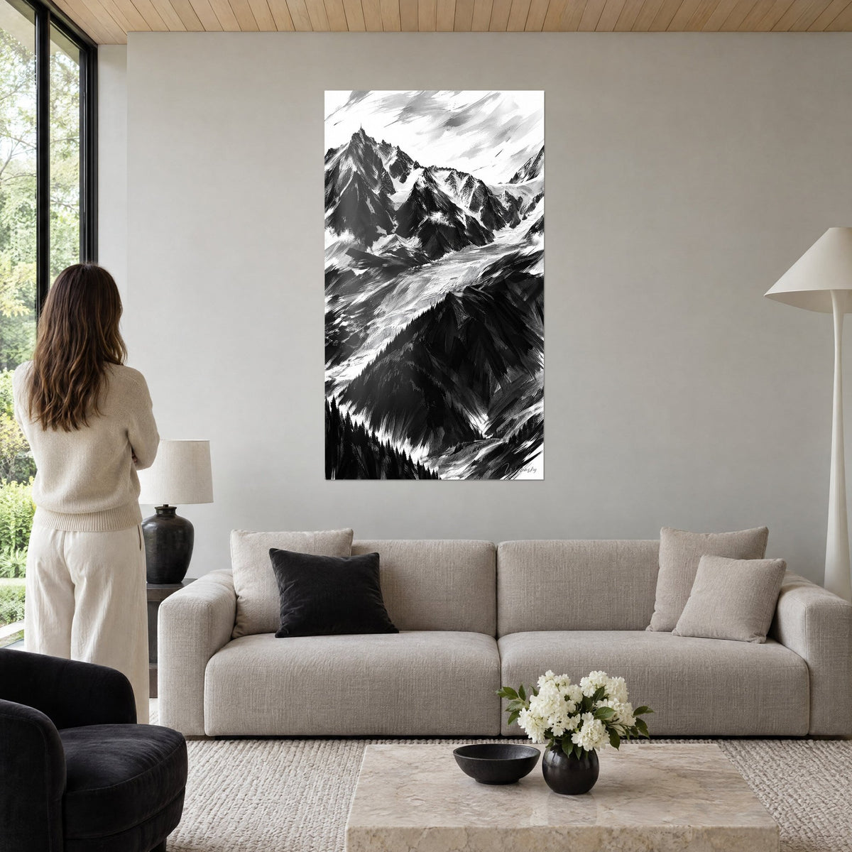 Schilderij gletsjer in zwart-wit, steile zwarte sparren en besneeuwde kammen, vrouw van achteren, boven een bank in een lichte woonkamer