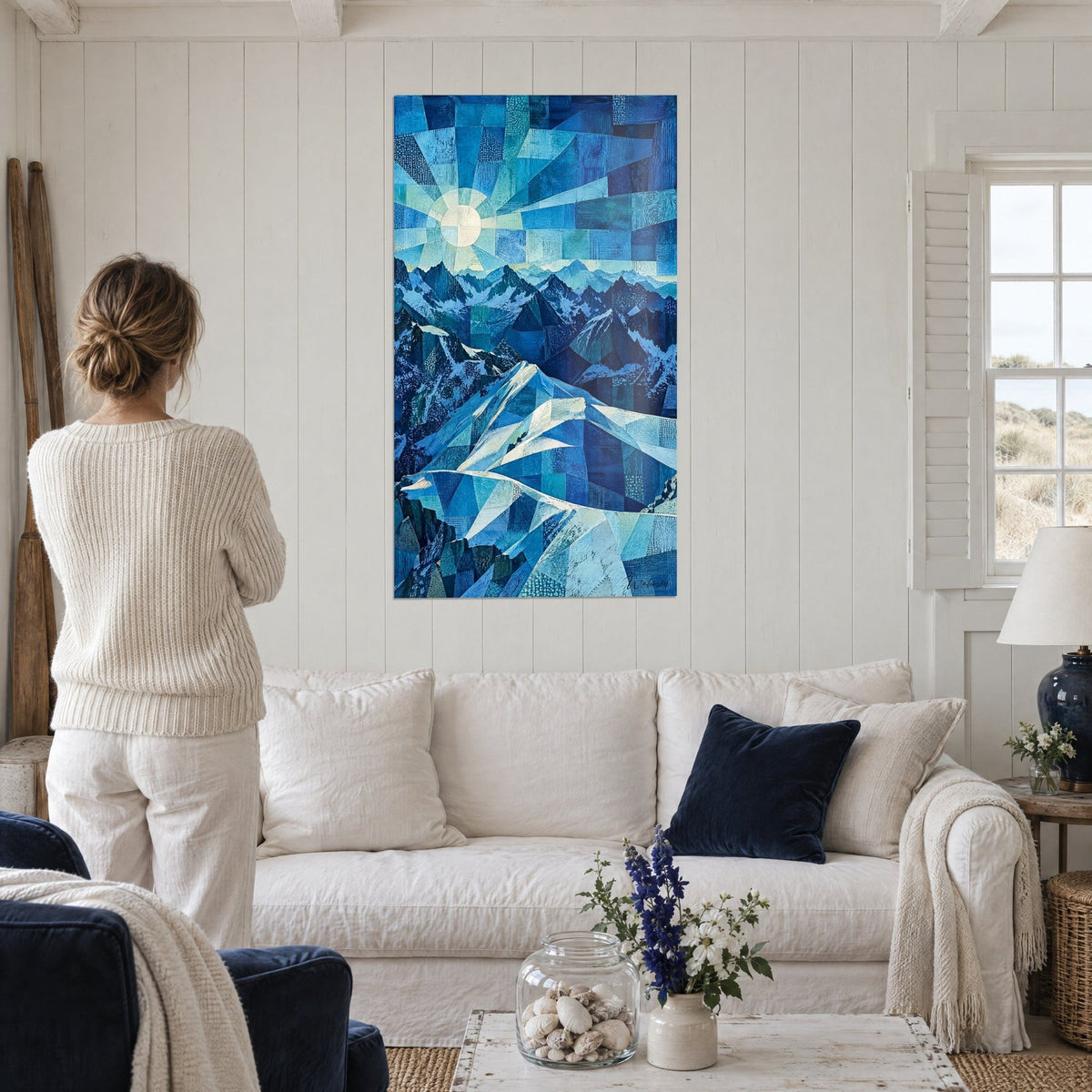 Schilderij ivoorkleurige zon met waaiervormige stralen boven gefacetteerde blauwe kammen, vrouw van achteren, boven een bank in een lichte woonkamer