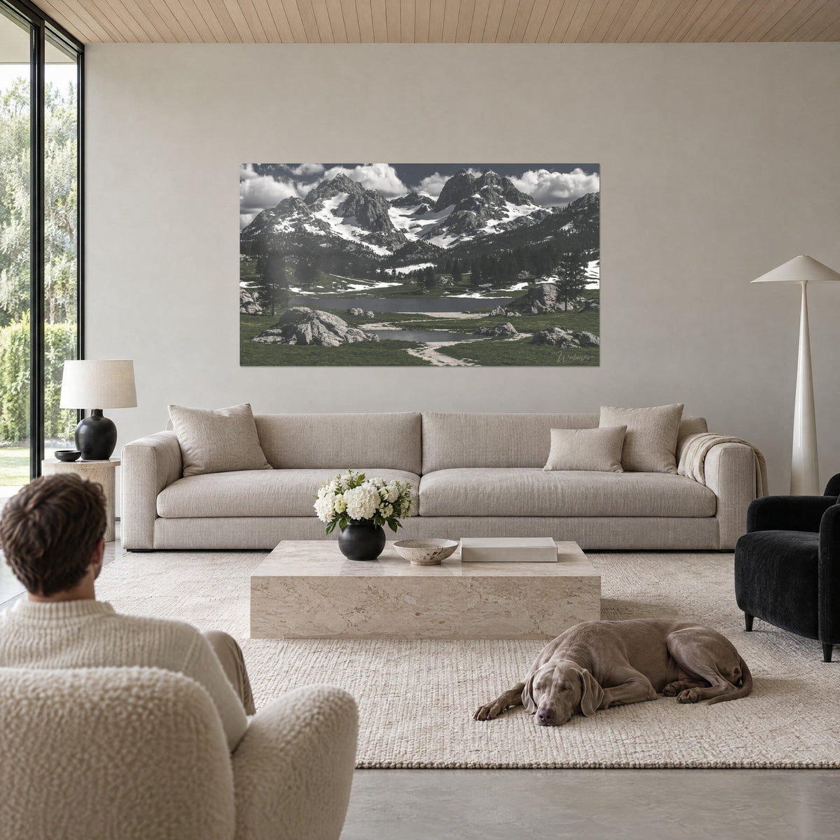 Schilderij besneeuwde rotstoppen die boven een grijs meer uittorenen, mosgroene weide, man van achteren en Weimaraner, boven een bank in een lichte woonkamer