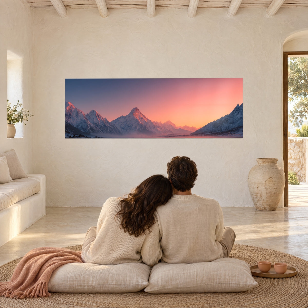 Cuadro paisaje de alta montaña teñido de coral e índigo, contemplado por una pareja sentada sobre cojines blancos