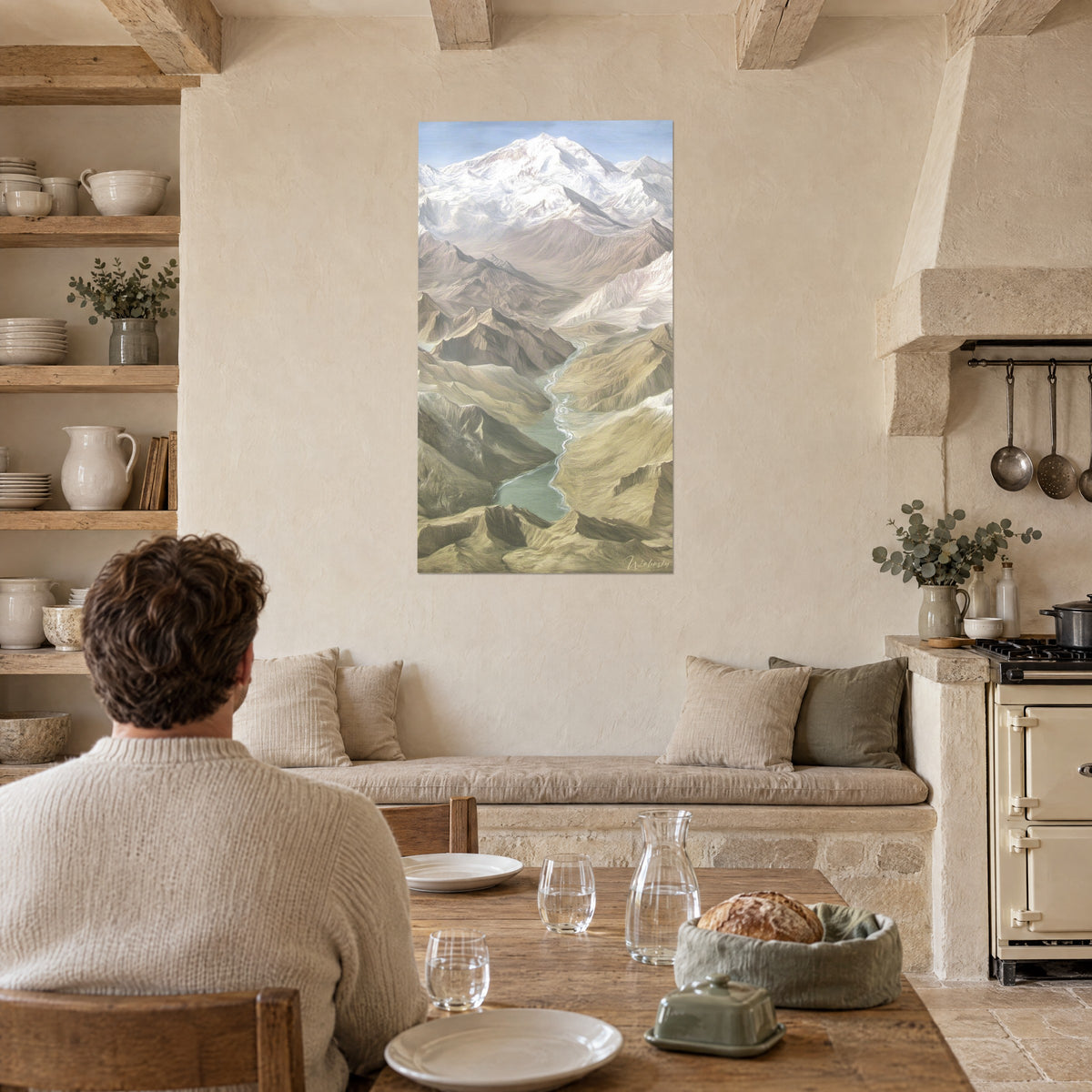 Tableau panorama de vallée aux arêtes brunes, sommet givré et lac vert pâle ; homme de dos attablé dans une cuisine en pierre claire
