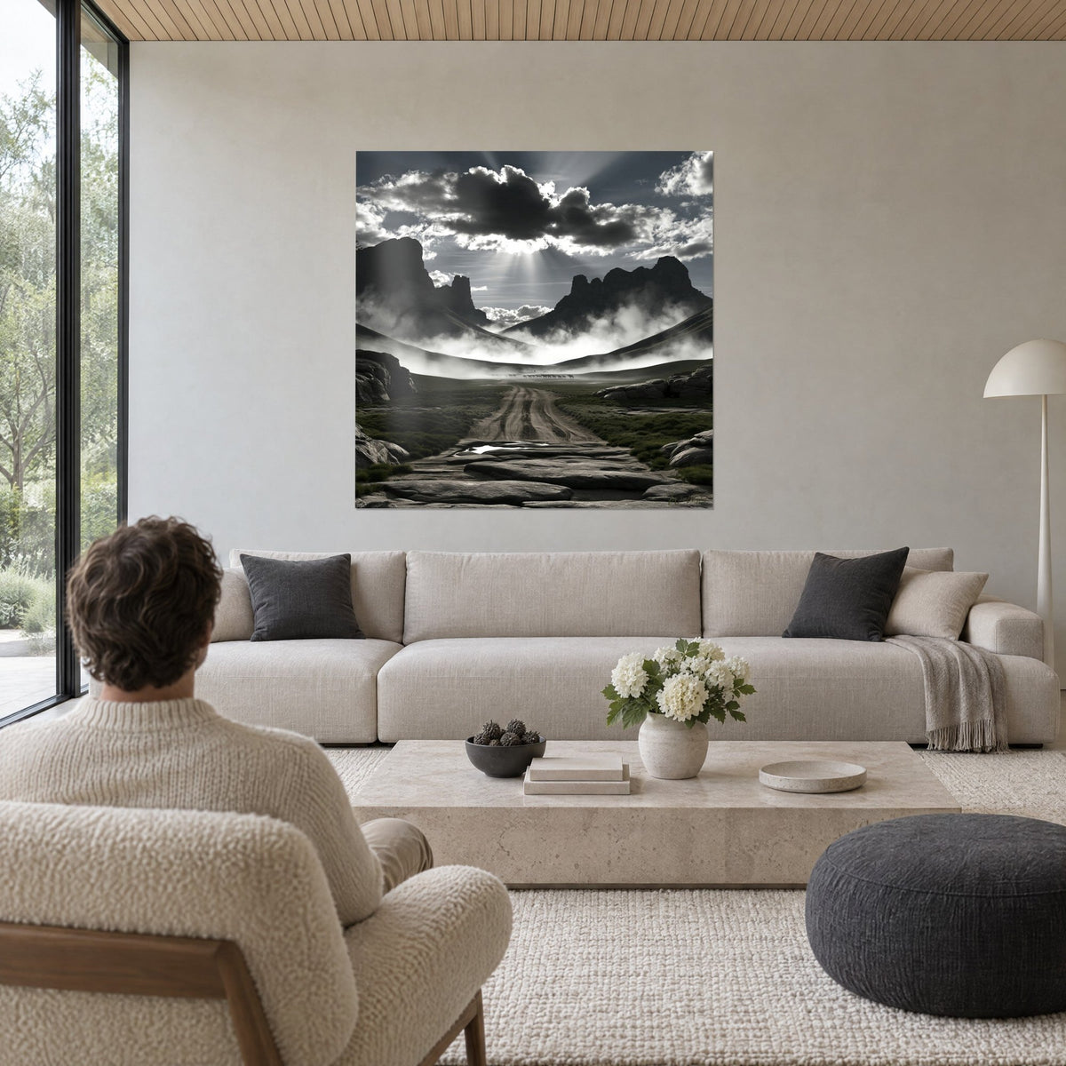 Wall art stone path leading to a misty valley, black and white sky, rays of light, man seen from behind above a sofa in a bright living room
