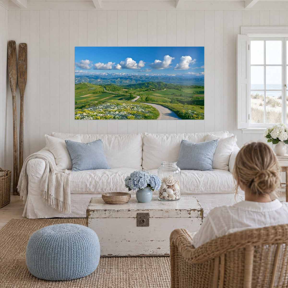 Tableau route sinueuse grise entre collines vertes, nuages blancs sur ciel bleu, au-dessus d'un canapé dans un salon lumineux