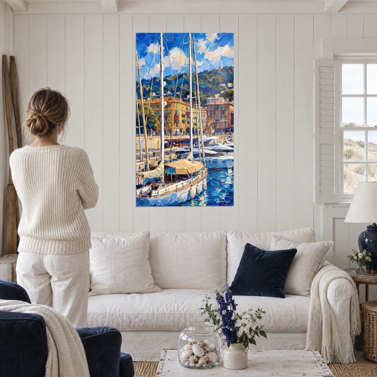 Schilderij zeilboten afgemeerd naast witte masten, okerkleurige gevels en blauwe lucht, vrouw van achteren boven een bank in een lichte woonkamer