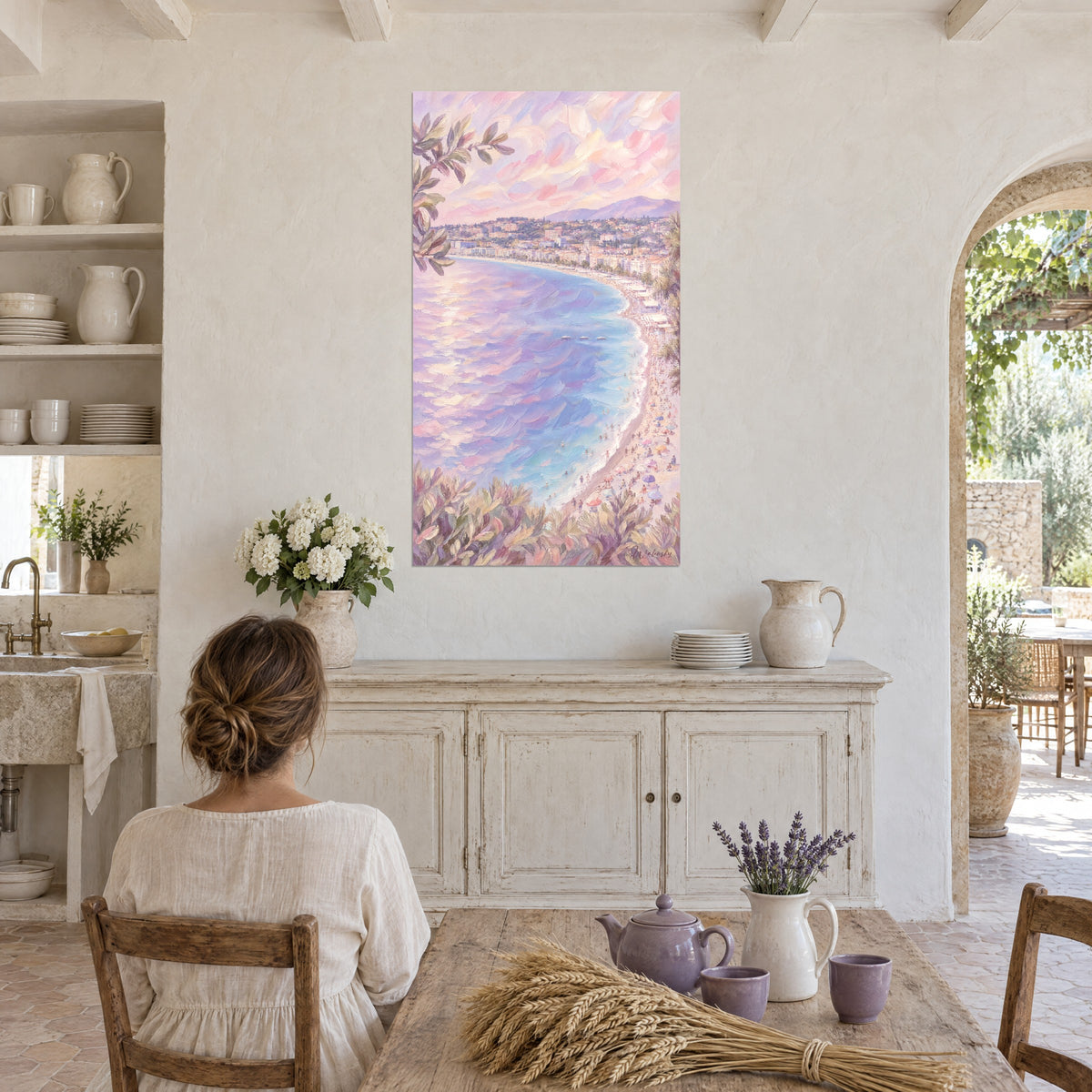 Quadro spiaggia di ciottoli con ombrelloni colorati, onde viola e cielo rosa cipria, una donna di spalle seduta a un tavolo di legno