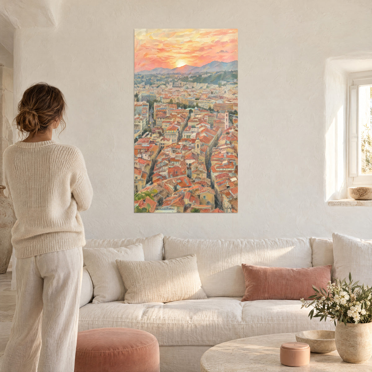 Schilderij panorama van terracotta dakpannen onder een roze en oranje lucht, vrouw van achteren, boven een bank in een lichte woonkamer