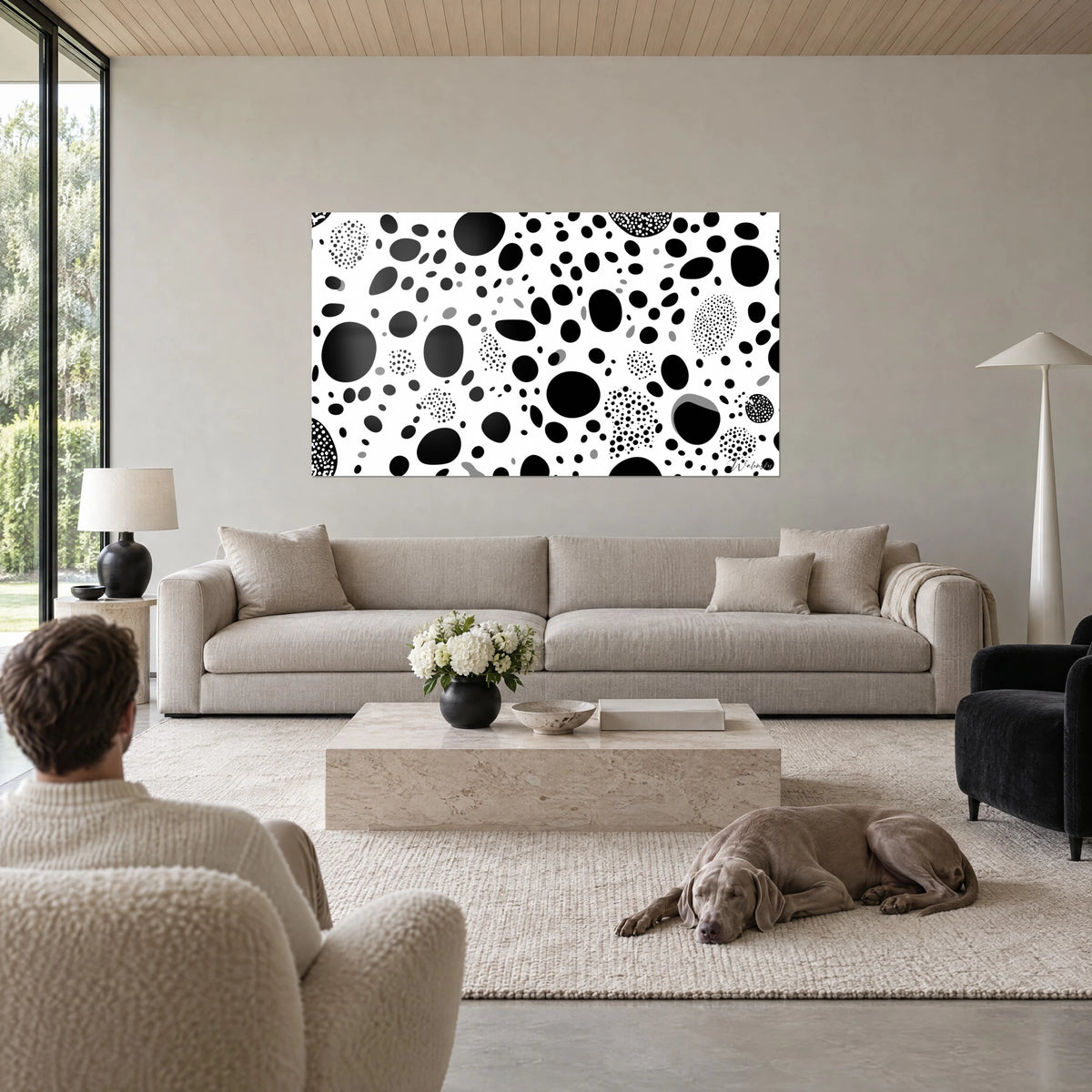 Wall art with black round spots on white background, clusters of dots and grey ovals, above a sofa in a bright living room