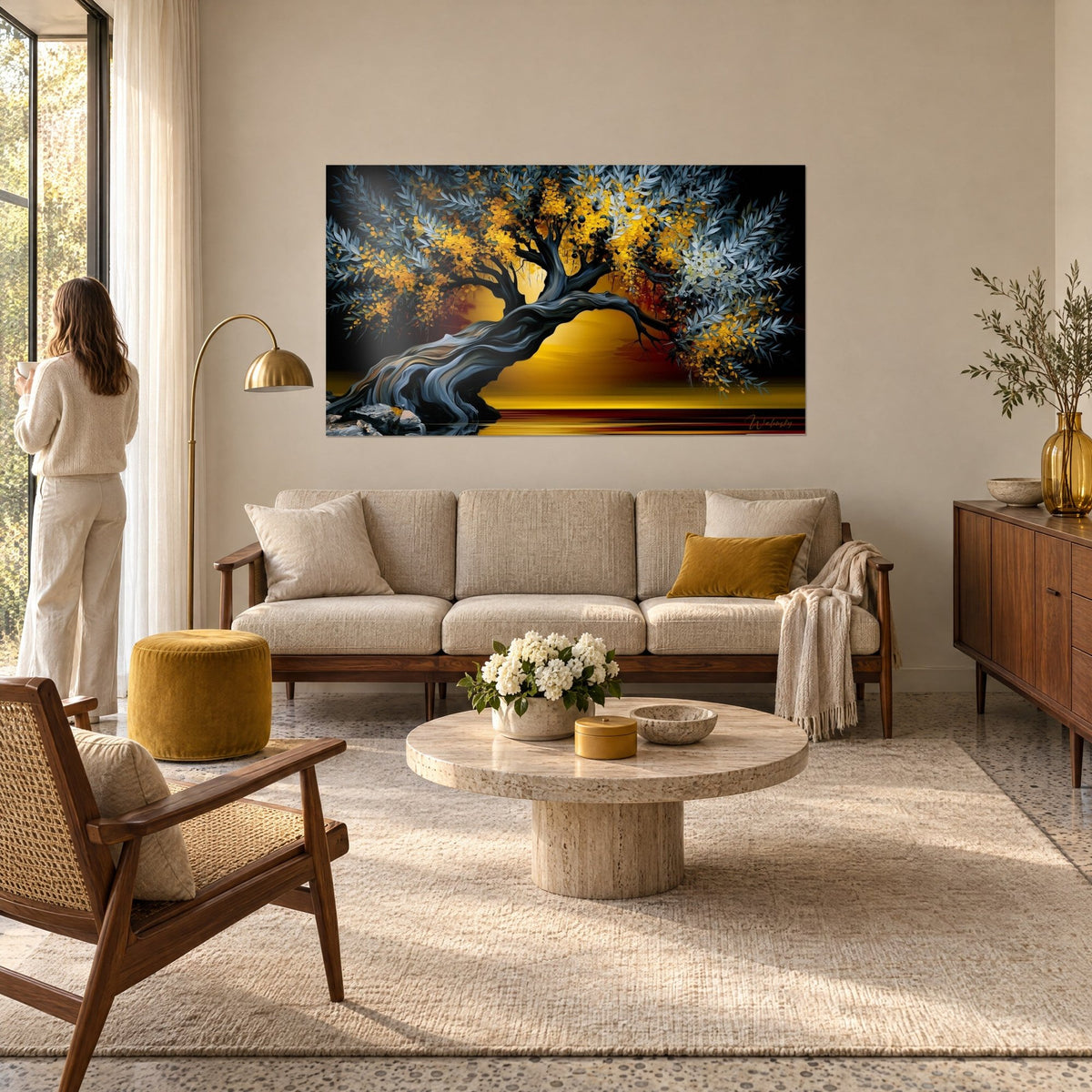 Wall art of an arched olive tree with grey-blue foliage and golden petals on a black background, woman seen from behind, above a sofa in a bright living room