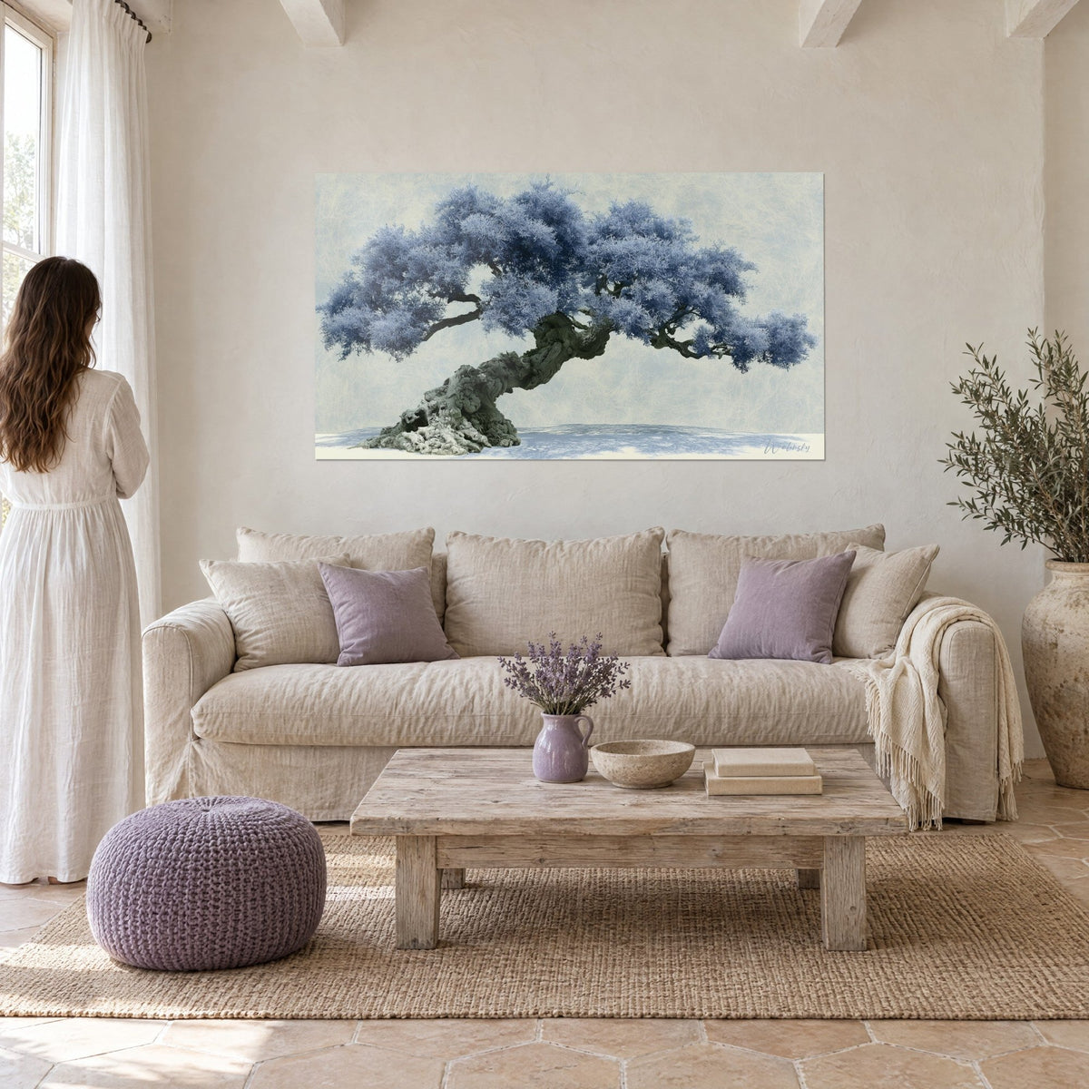 Wall art of a centuries-old olive tree with frosted blue foliage and a gnarled grey trunk on a cream background, above a sofa in a bright living room