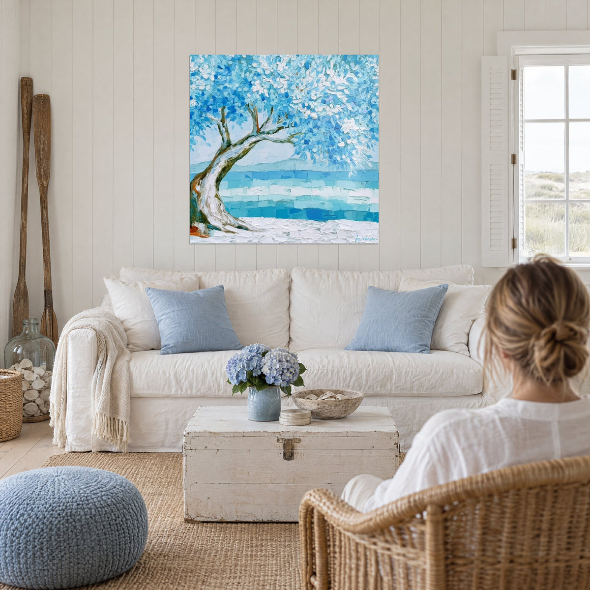 Quadro di un ulivo piegato dalle foglie azzurro cielo e bianche di fronte a un mare turchese, sopra un divano in un soggiorno luminoso