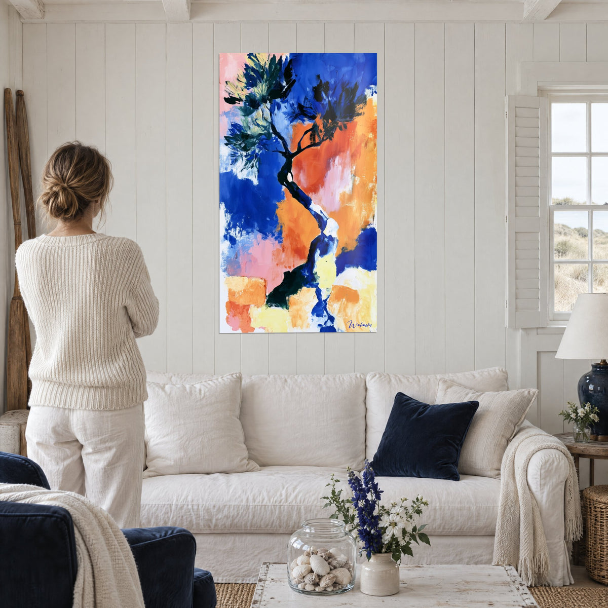 Wall art olive tree with a winding cobalt blue trunk and bright orange accents, black foliage, person seen from behind, above a sofa in a bright living room
