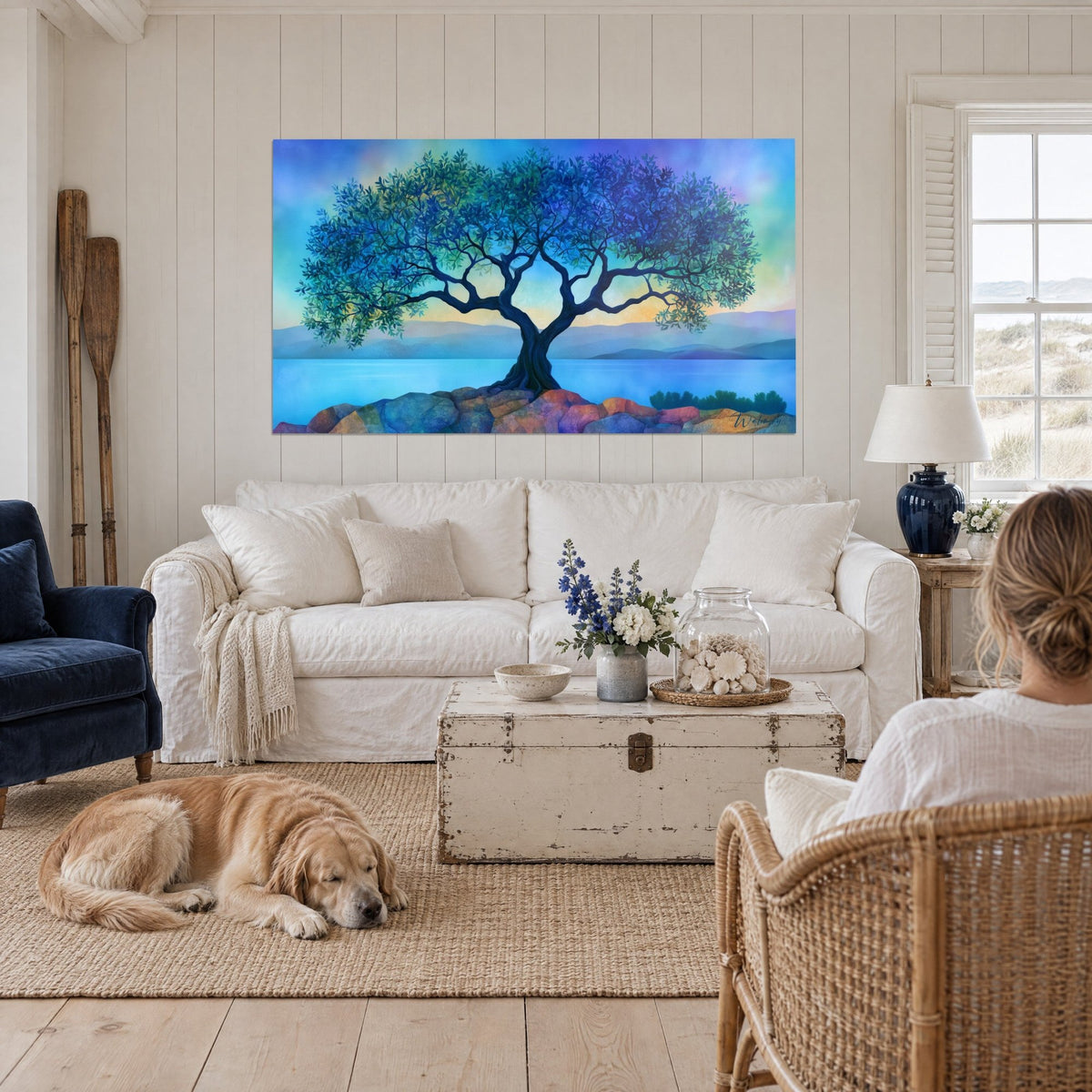 Wall art of a gnarled olive tree with midnight blue and turquoise foliage on ochre rocks, a reclining retriever, above a sofa in a bright living room