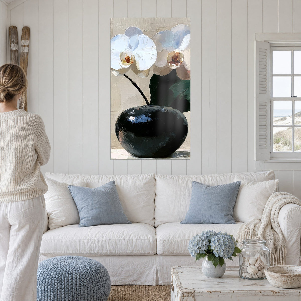 Wall art white orchids with ochre hearts in a round black vase, dark green leaf, cream wall, above a sofa in a bright living room